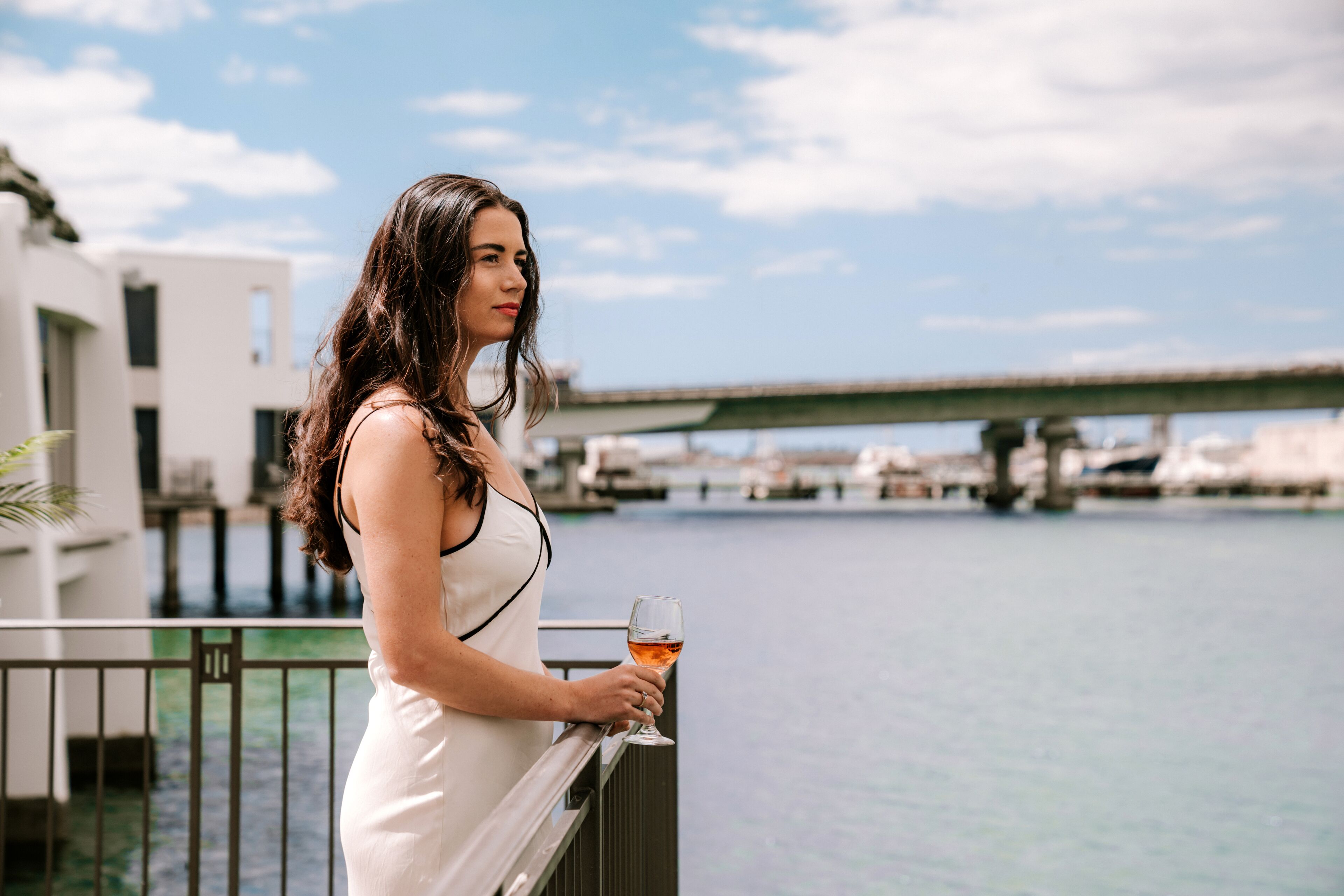 Eine Frau in einem weißen Kleid steht auf einem Balkon mit einem Weinglas und blickt auf eine Marina mit Booten und einer Brücke im Hintergrund.