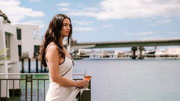 Eine Frau in einem weißen Kleid steht auf einem Balkon mit einem Weinglas und blickt auf eine Marina mit Booten und einer Brücke im Hintergrund.