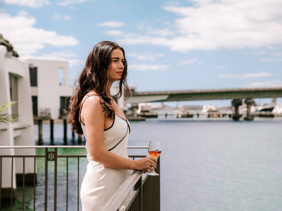 Eine Frau in einem weißen Kleid steht auf einem Balkon mit einem Weinglas und blickt auf eine Marina mit Booten und einer Brücke im Hintergrund.