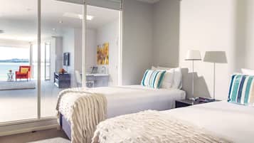 A hotel room with two beds, a desk, and a chair. There is a view of the sea through large windows.