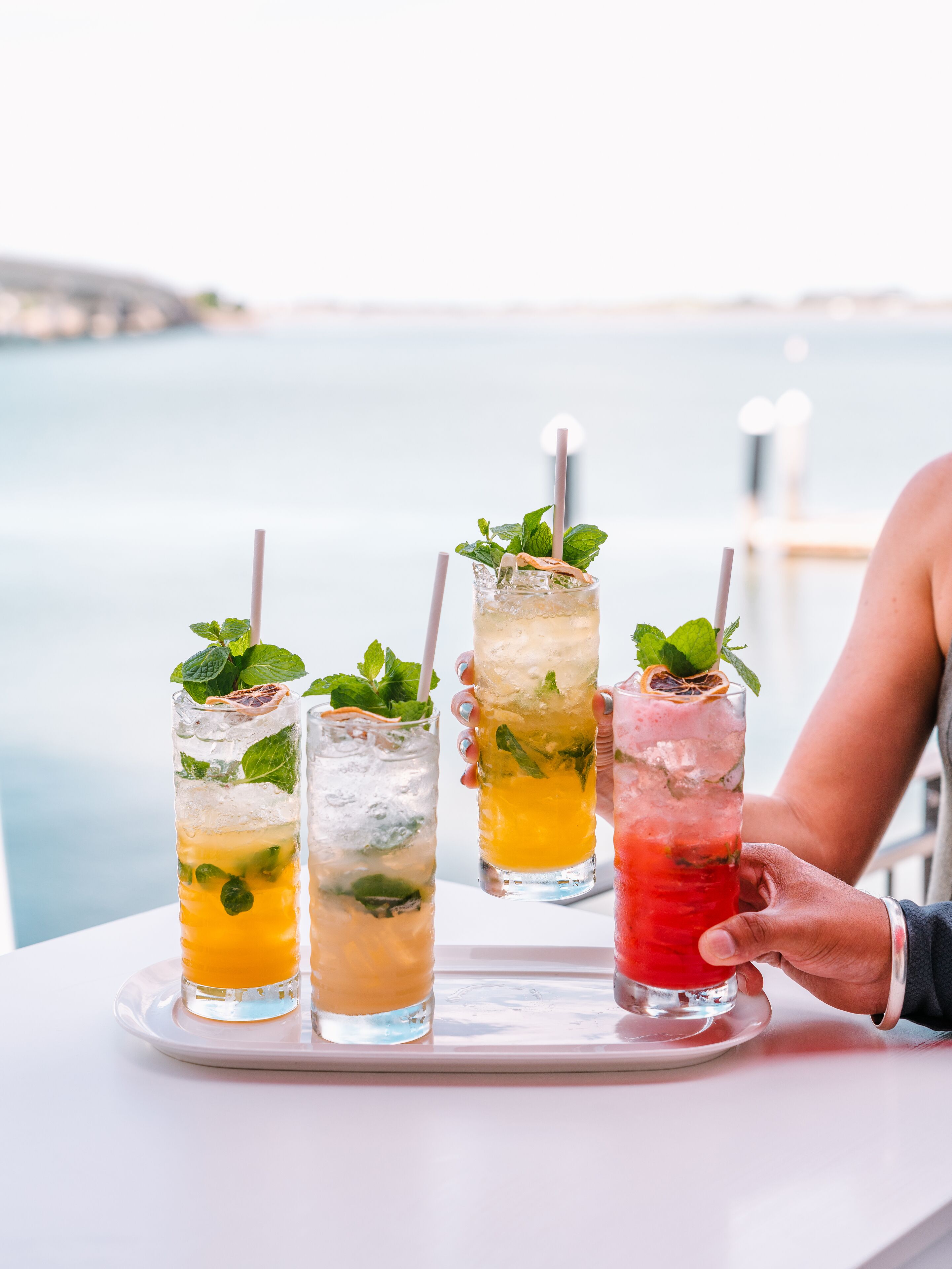 Four tall glasses of iced drinks with mint leaves and straws, placed on a tray.