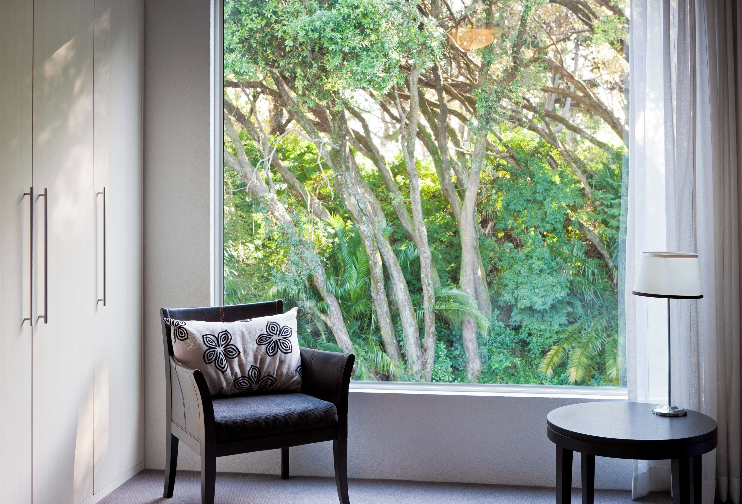 A room with a view of a lush garden through a large window, a black chair with a patterned pillow, and a small round table with a lamp.
