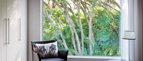 A room with a view of a lush garden through a large window, a black chair with a patterned pillow, and a small round table with a lamp.