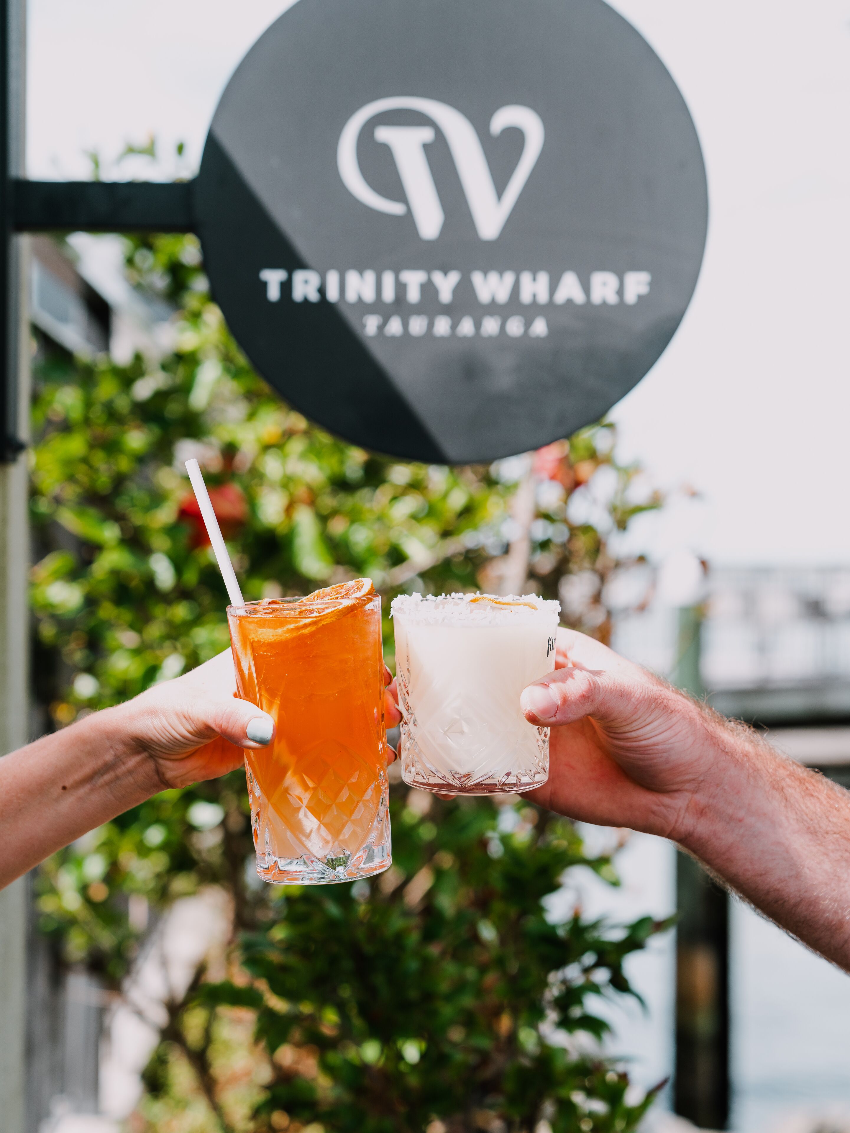 Two hands holding glasses of beverages, with a sign reading "TRINITY WHARF" in the background.