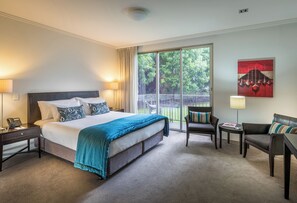 A hotel room with a large bed, a sitting area with a chair and a small table, a lamp, and a view of the outdoors through a sliding door. - Trinity Wharf Tauranga (Tauranga)