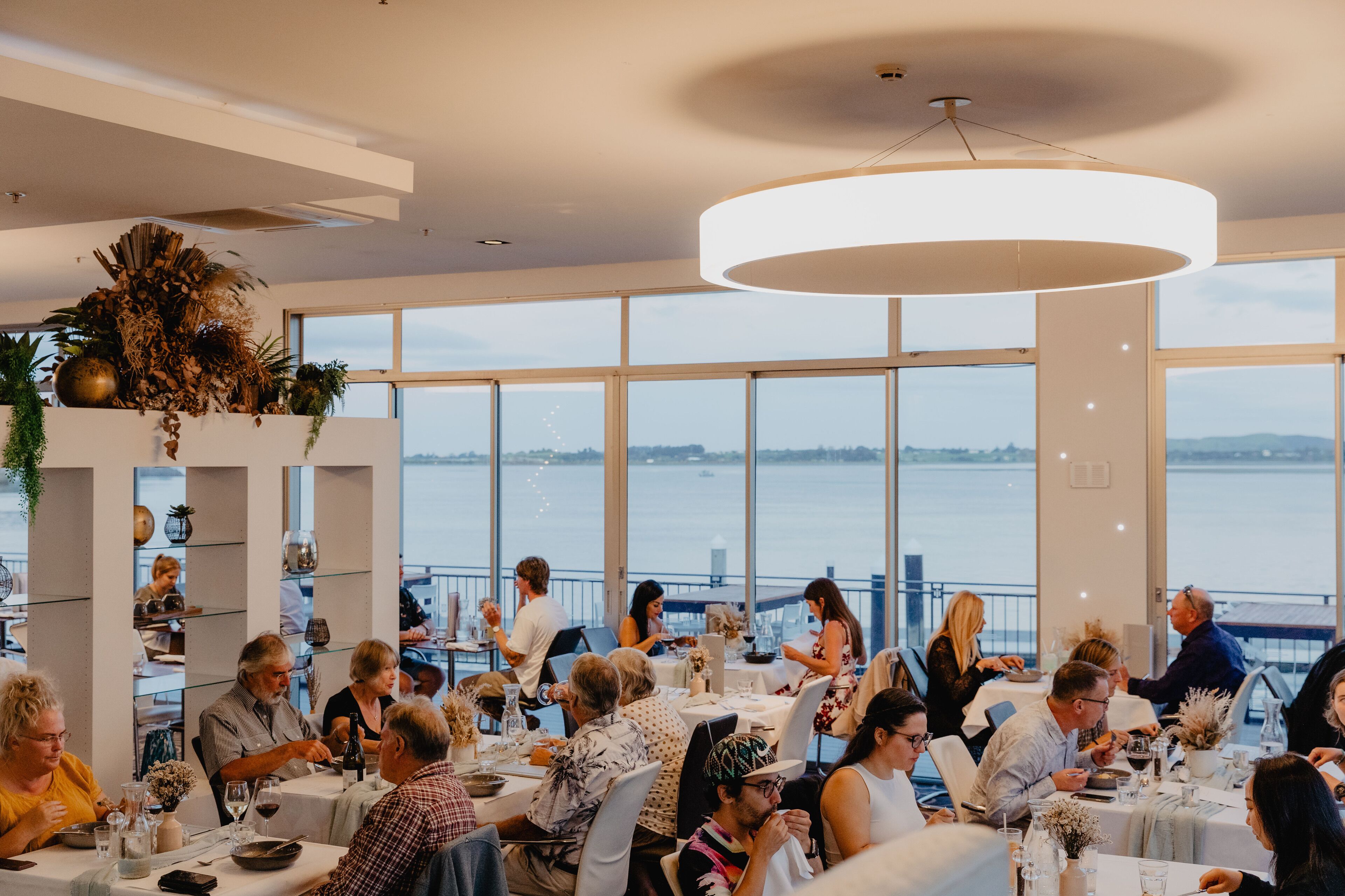Un restaurante con amplios ventanales que ofrecen vistas al agua, diseño interior moderno y una variedad de mesas para comer.