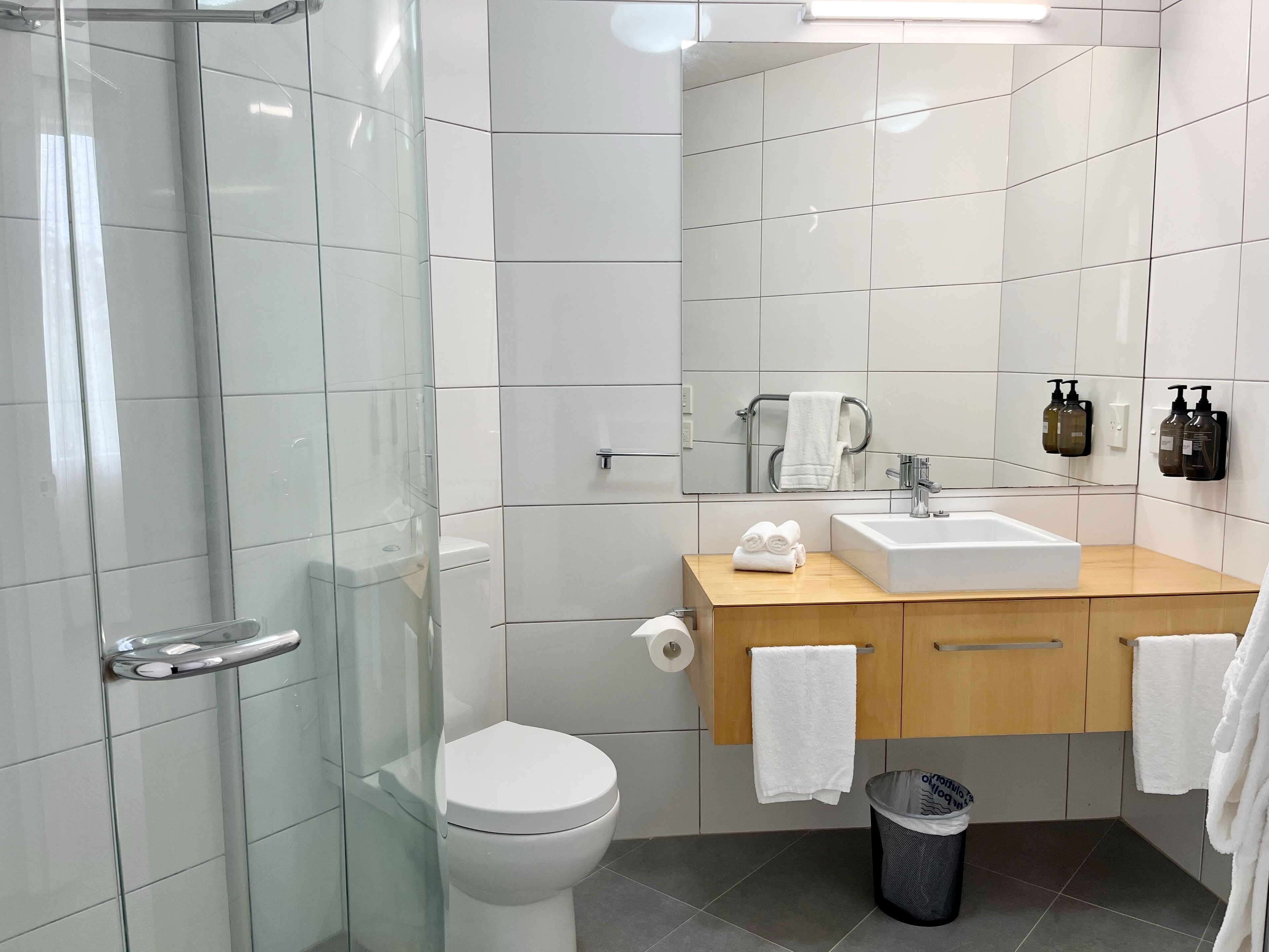 Un baño moderno con cabina de ducha de vidrio, inodoro blanco, lavabo con tocador de madera y espejo.