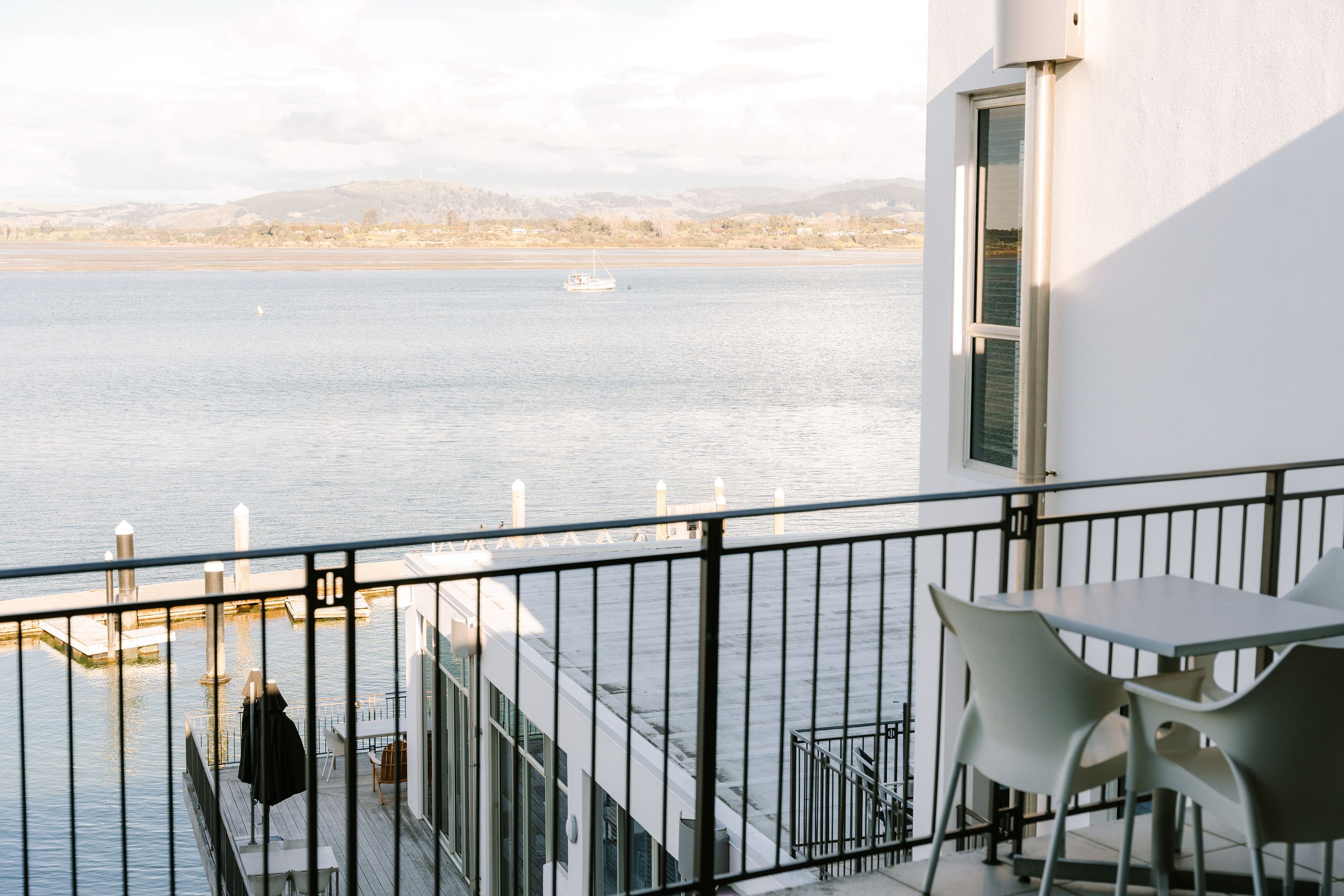 Ein Balkon mit Tisch und Stühlen, der auf ein ruhiges Gewässer mit Uferlinie und Hügeln blickt.