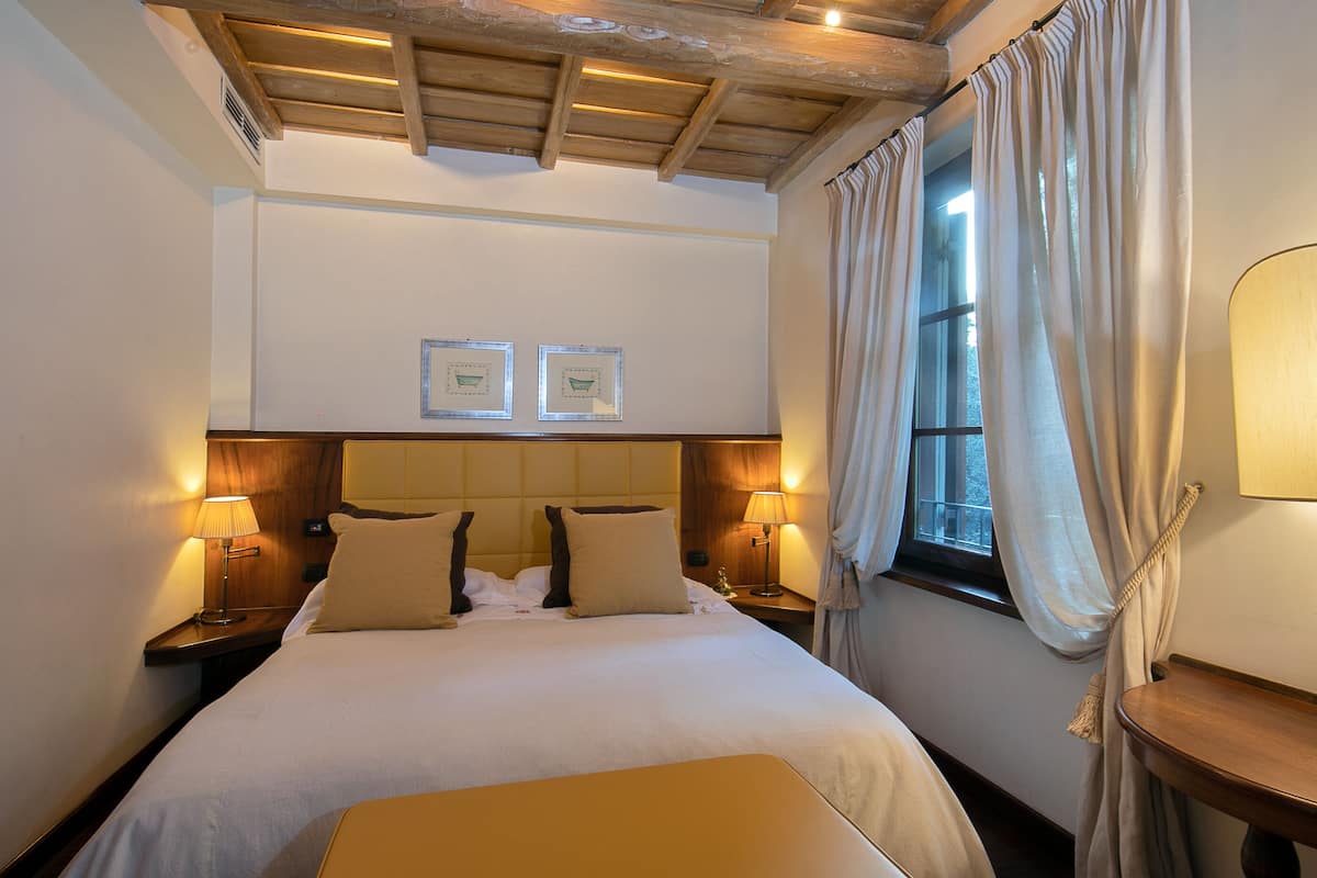 A hotel room with a large bed, two bedside lamps, a wooden headboard, a window with curtains, and a small table with a lamp.