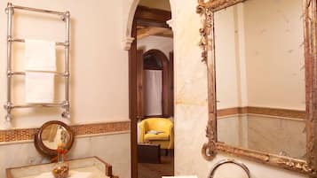 A bathroom with a marble sink, wooden vanity, a large ornate mirror, and a yellow chair in the background.