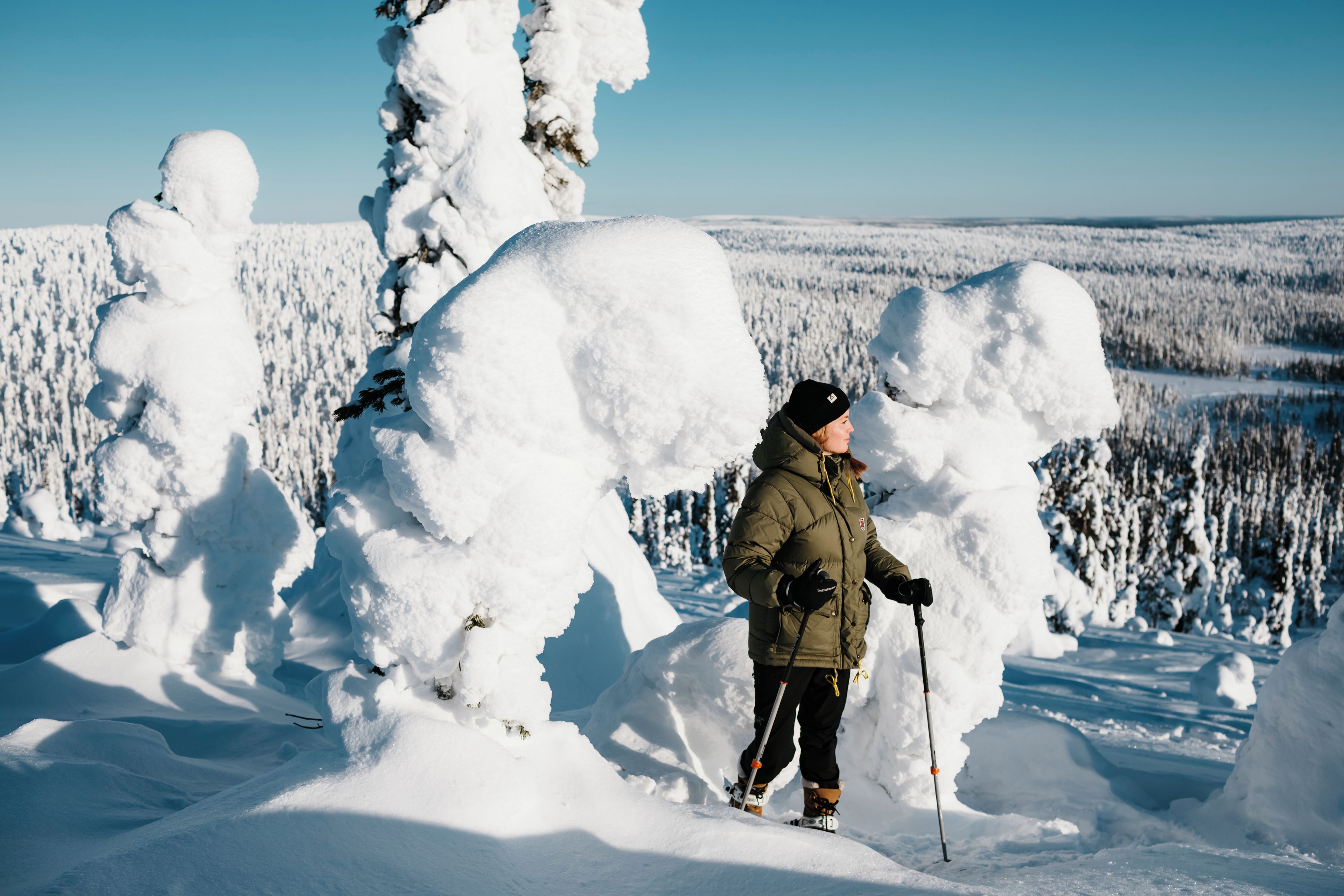 Henkilö seisoo lumisessa maisemassa, jonka taustalla on lumipeitteisiä puita ja kirkas taivas.