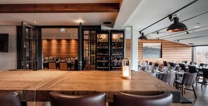 Restaurante com uma longa mesa de madeira, cadeiras de couro e uma adega visível através de portas de vidro.