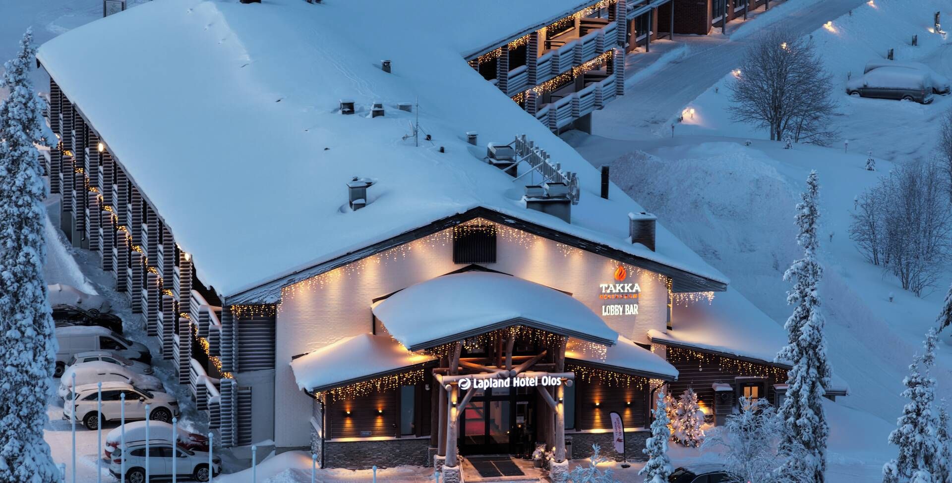 En snötäckt byggnad med en bar i lobbyn och en skylt med texten ”Tapland Hotel Olmos”.