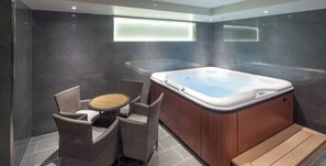 A modern bathroom with a hot tub, wicker furniture, and dark tiled walls.