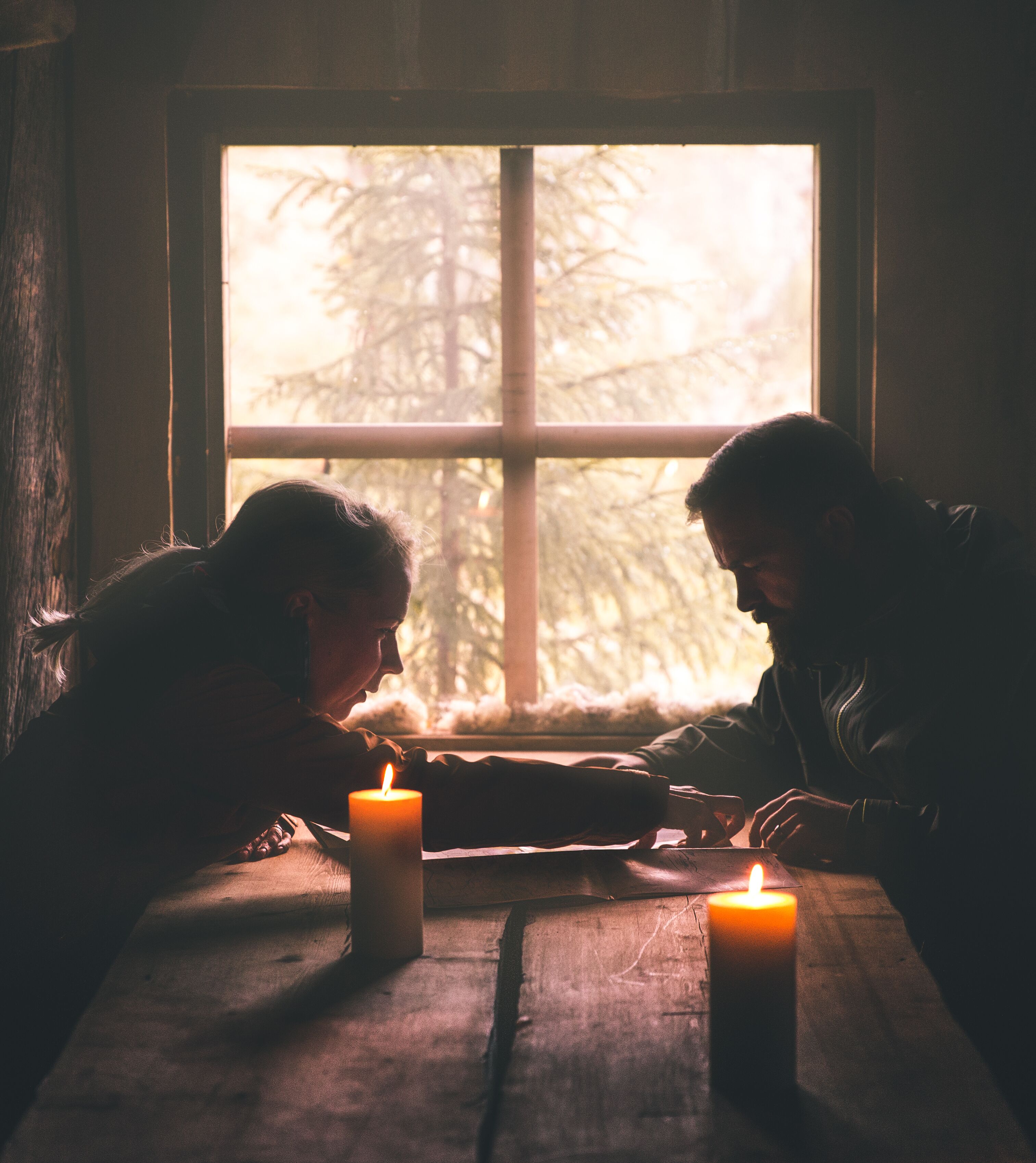 Two people sitting at a wooden table with two lit candles, in a room with a large window.