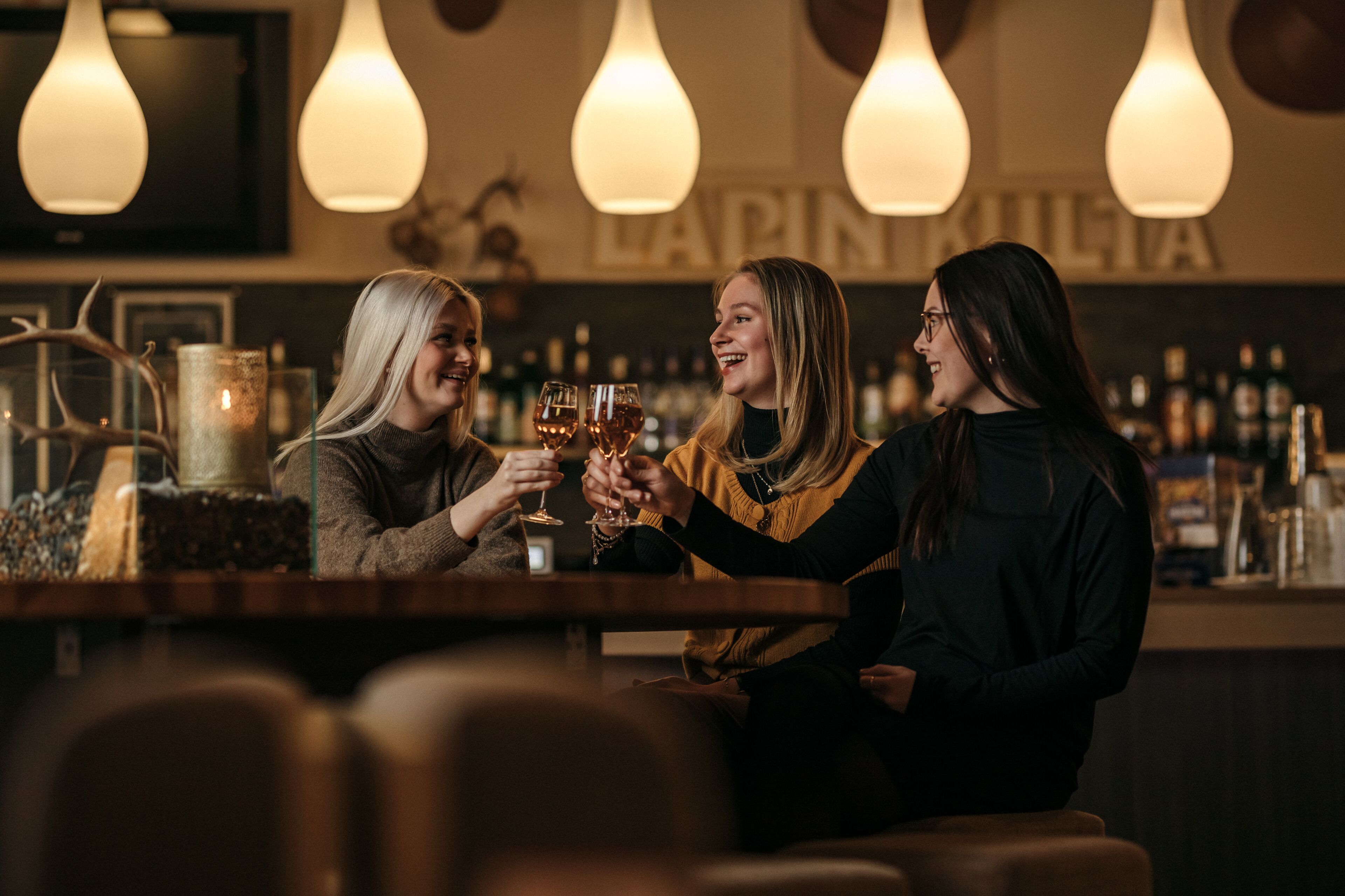 Three people toasting with drinks at a bar with hanging light fixtures and a visible sign that reads "LAPIN MILIA".