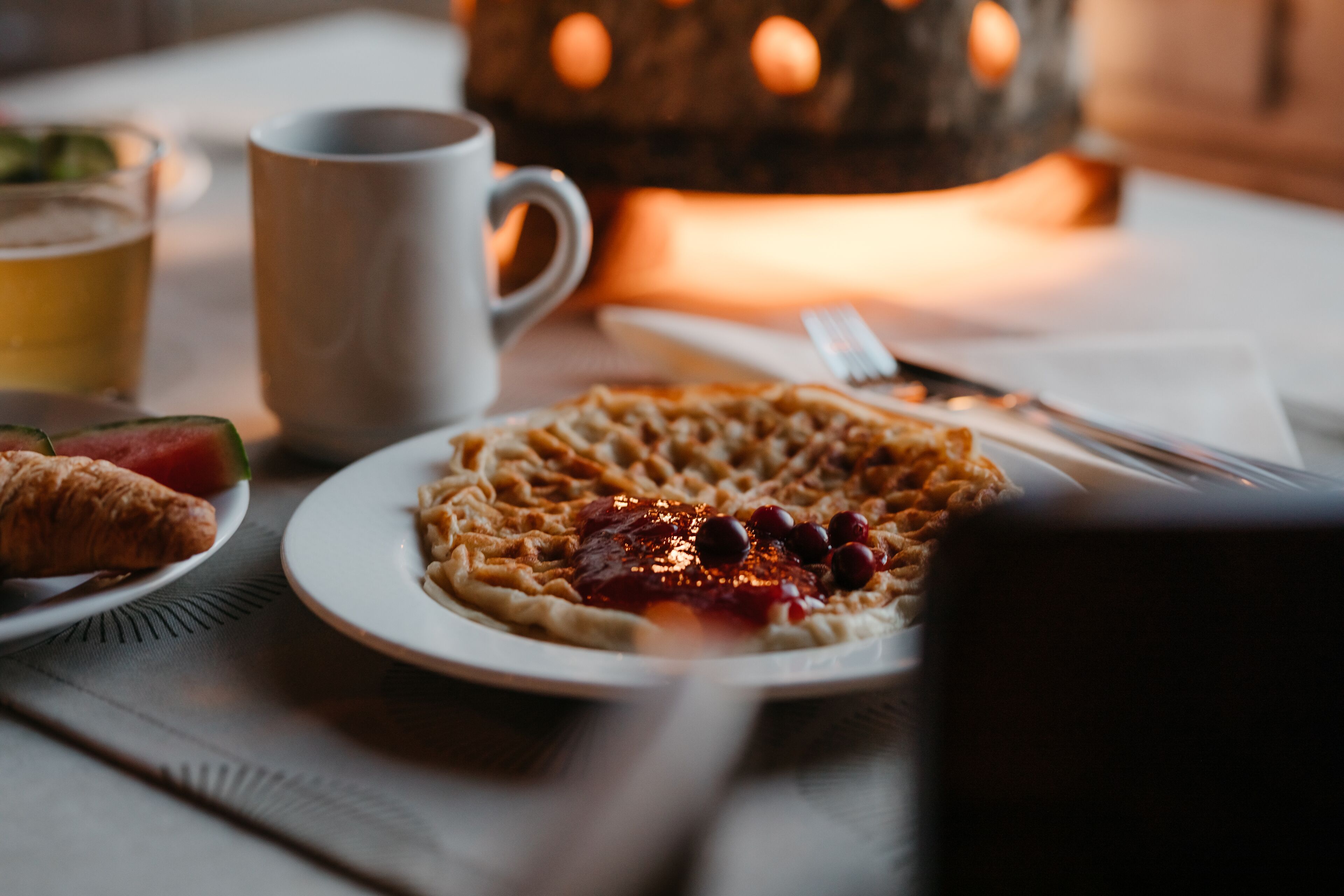 Um prato de waffles com calda e frutas vermelhas, um croissant, uma xícara de café e um copo de cerveja sobre uma mesa.