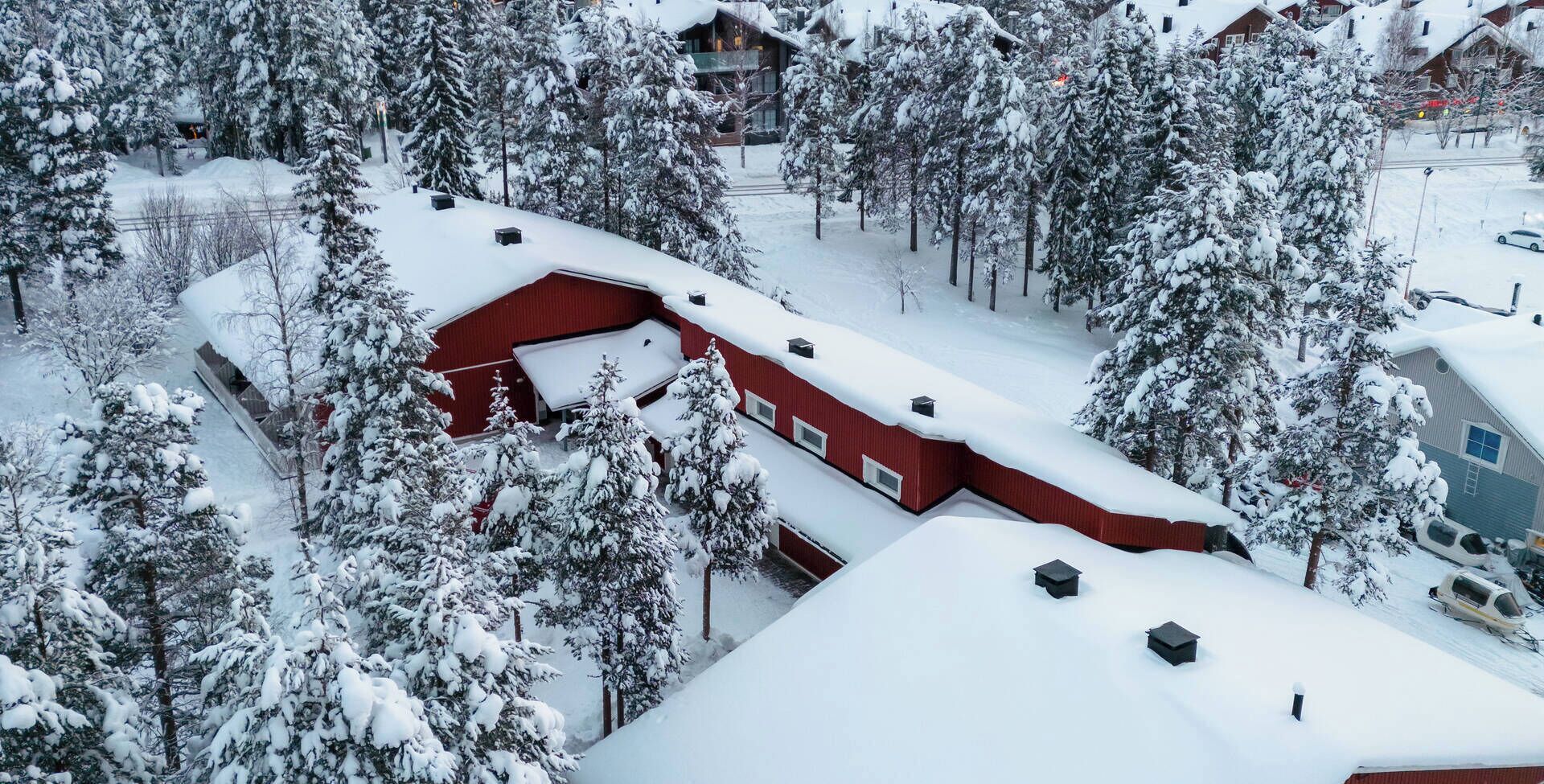 Un edificio rojo con varias ventanas y un tejado cubierto de nieve, rodeado de árboles cargados de nieve.