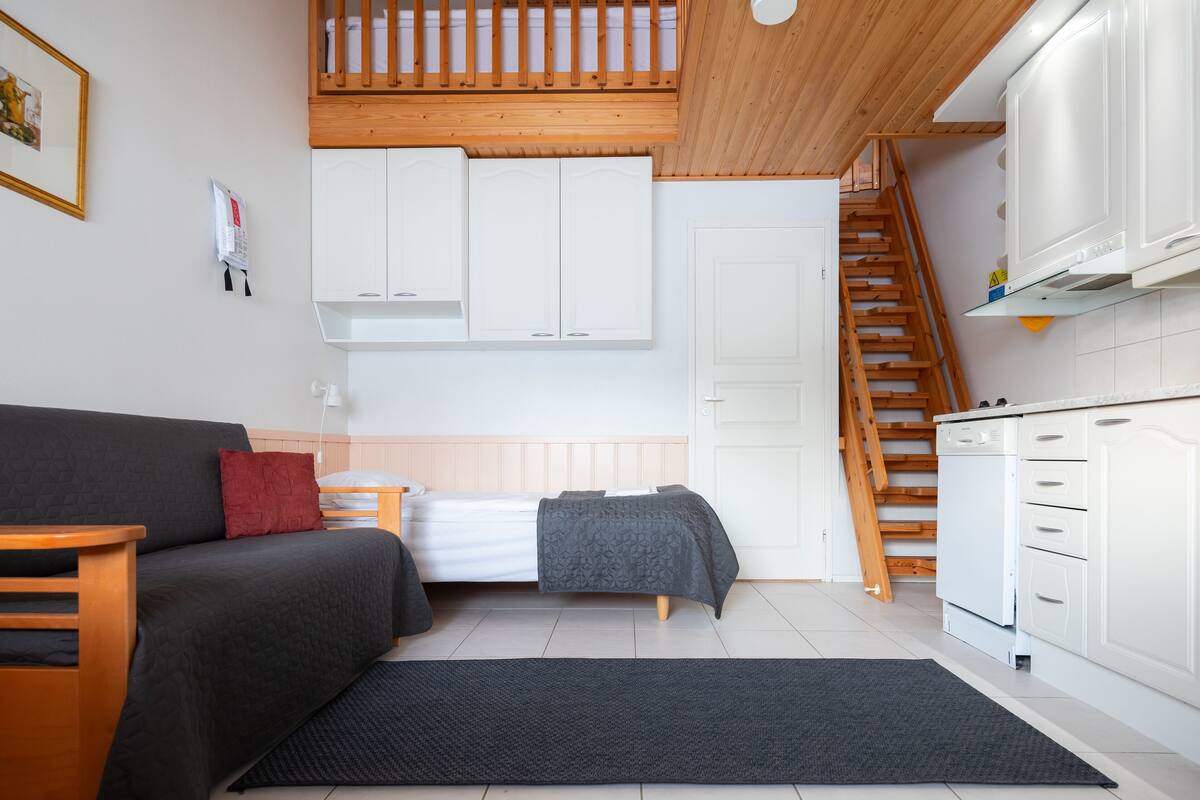 Een kleine woonruimte met een bed, een slaapbank, een keukenhoek en een trap.