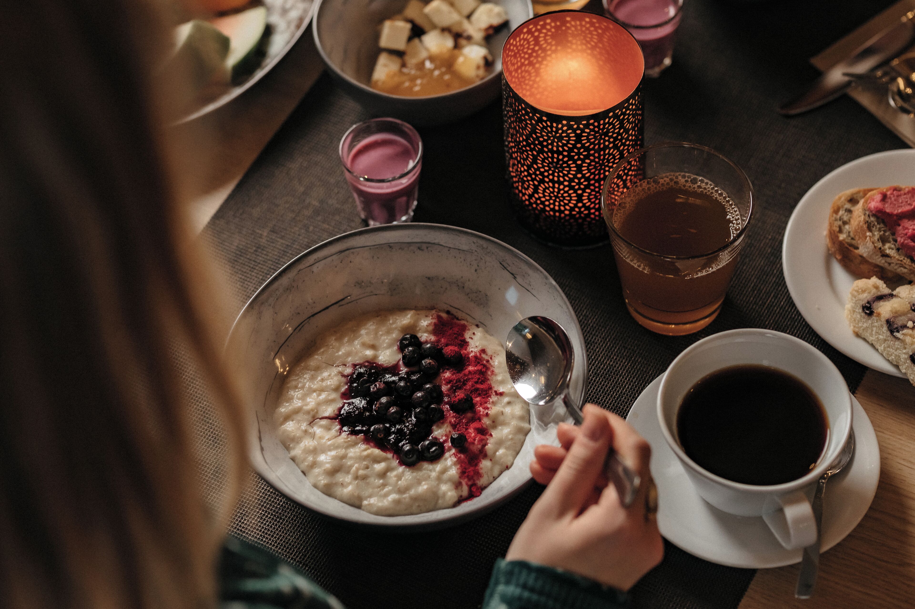 A dining table with a bowl of oatmeal topped with blueberries and red powder, a glass of tea, a cup of coffee, and a candle.