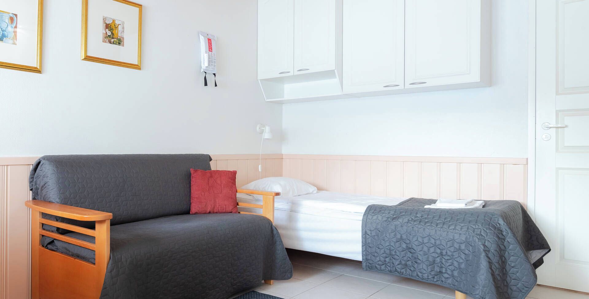 A small, neatly arranged room with a bed, a sofa, and a chair. There are framed pictures on the wall and a white cabinet above the bed.