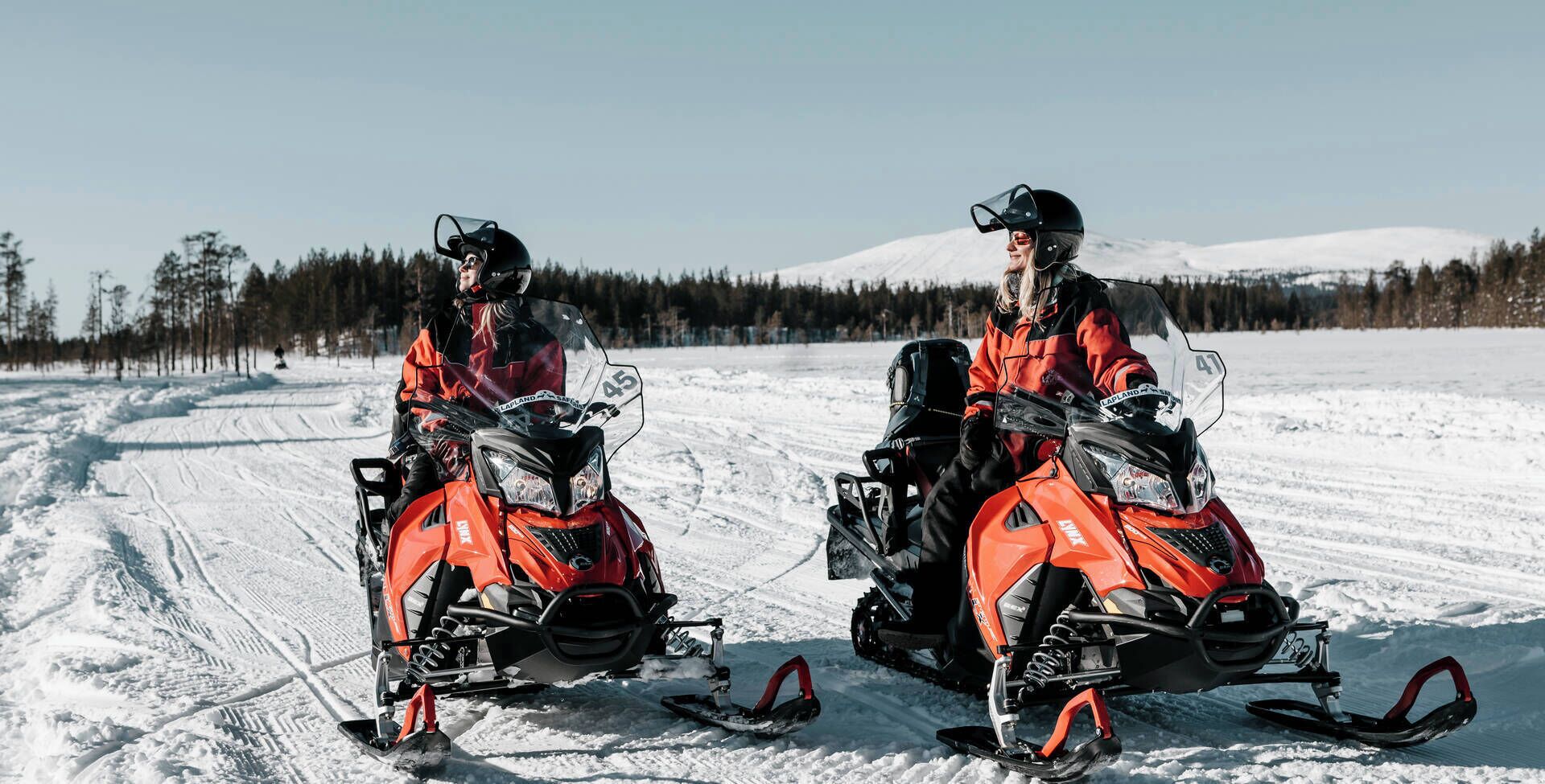 Two people on snowmobiles in a snowy landscape.