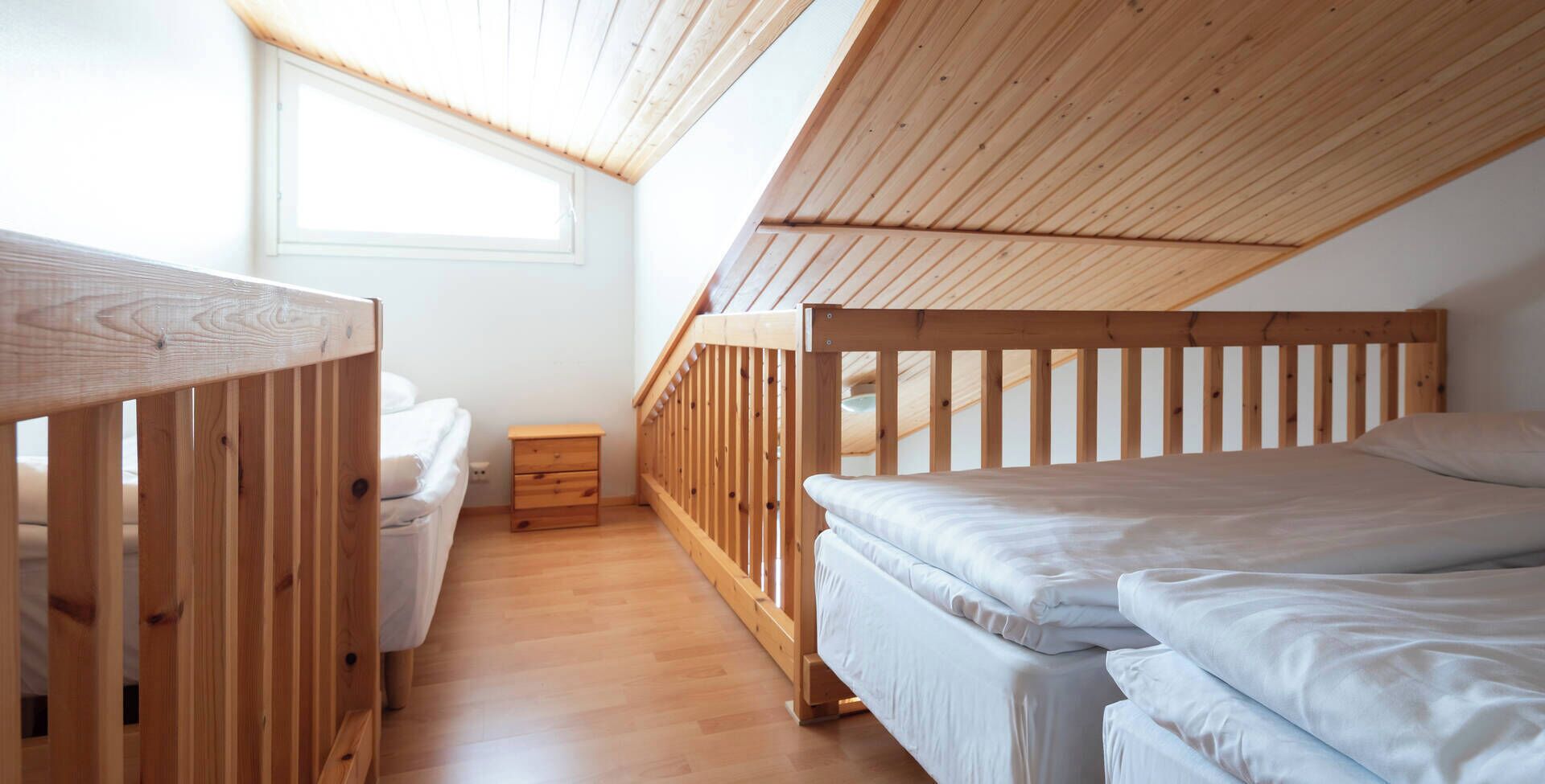 Un dormitorio con dos literas, piso de madera, una mesita de noche y una ventana que deja entrar luz natural.