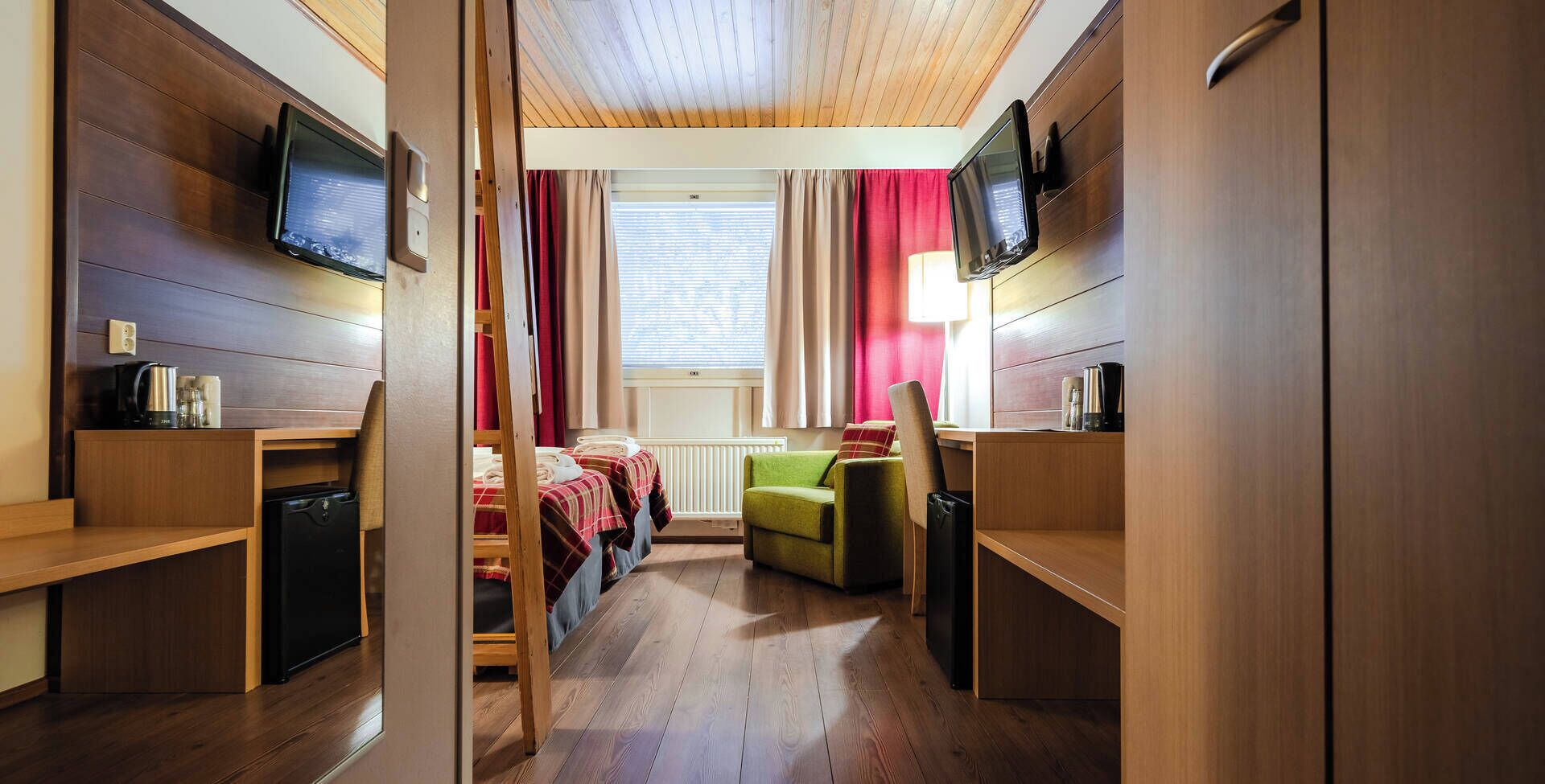 Une chambre d’hôtel compacte avec un plancher en bois, un lit avec une literie rouge et blanche, un fauteuil vert, un téléviseur fixé au mur et une fenêtre avec des rideaux.