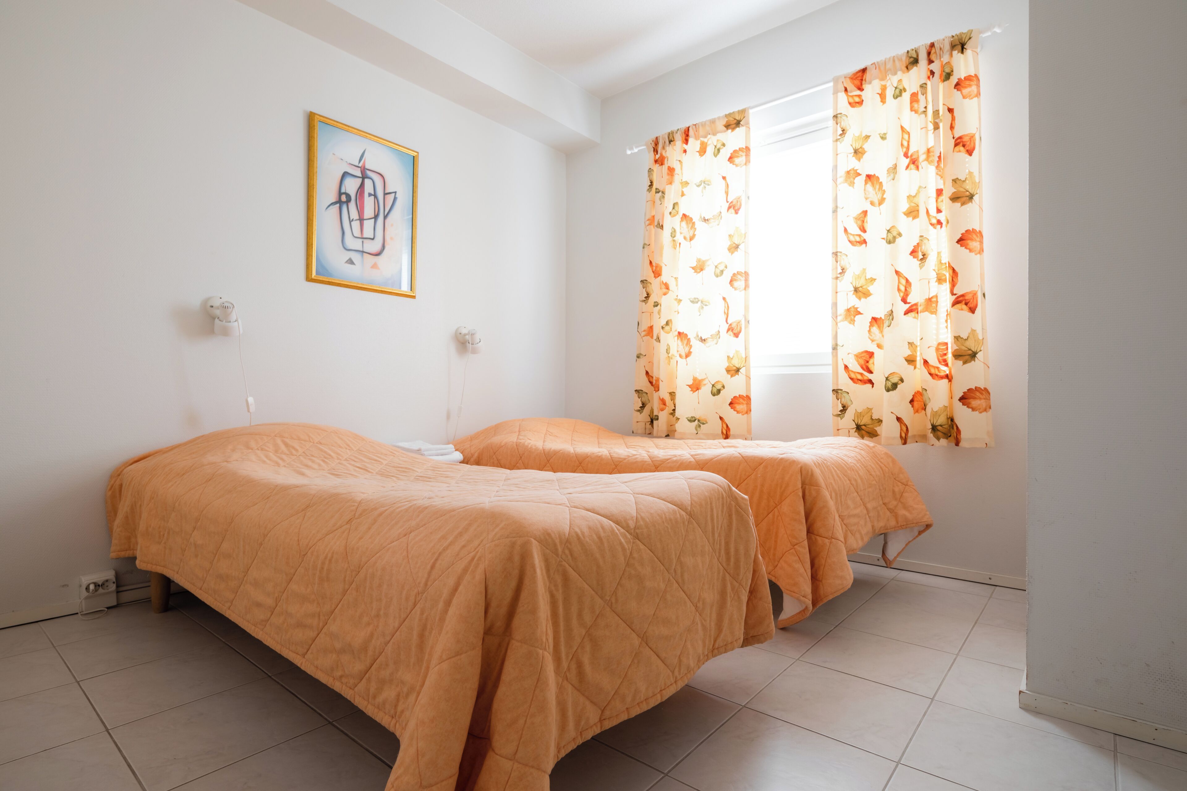 Une chambre avec deux lits, une fenêtre ornée de rideaux à motifs floraux, un tableau accroché au mur et un sol carrelé.