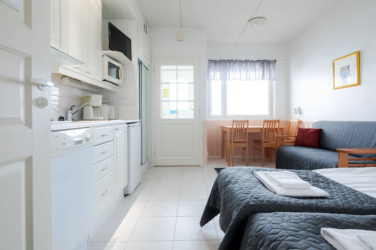 Een kleine kamer met een kitchenette, een bed met een gewatteerde spreidek, een eettafel met stoelen en een raam met gordijnen.