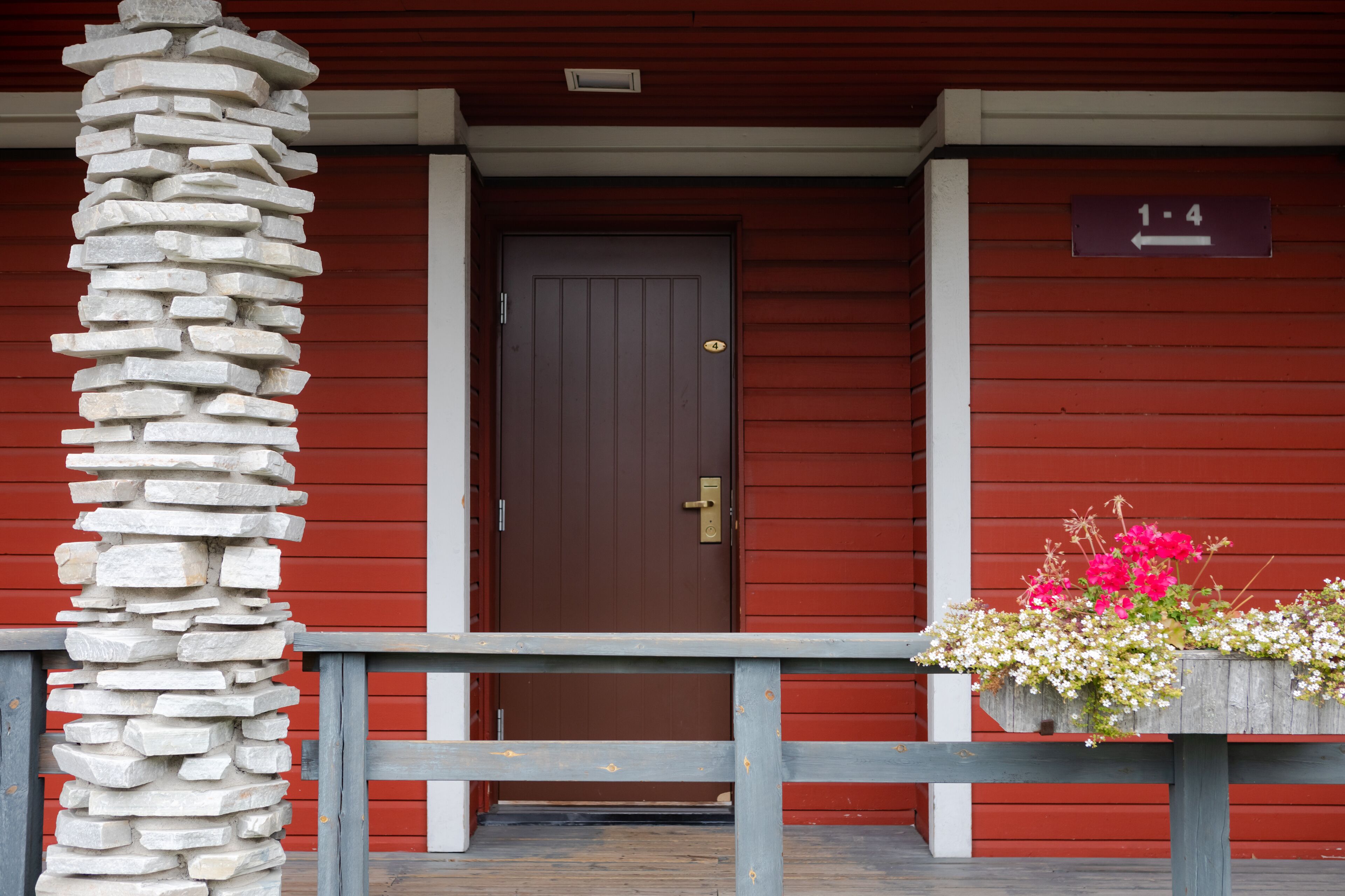 A red wooden building with a brown door, a stone pillar, and a flower planter on the porch.