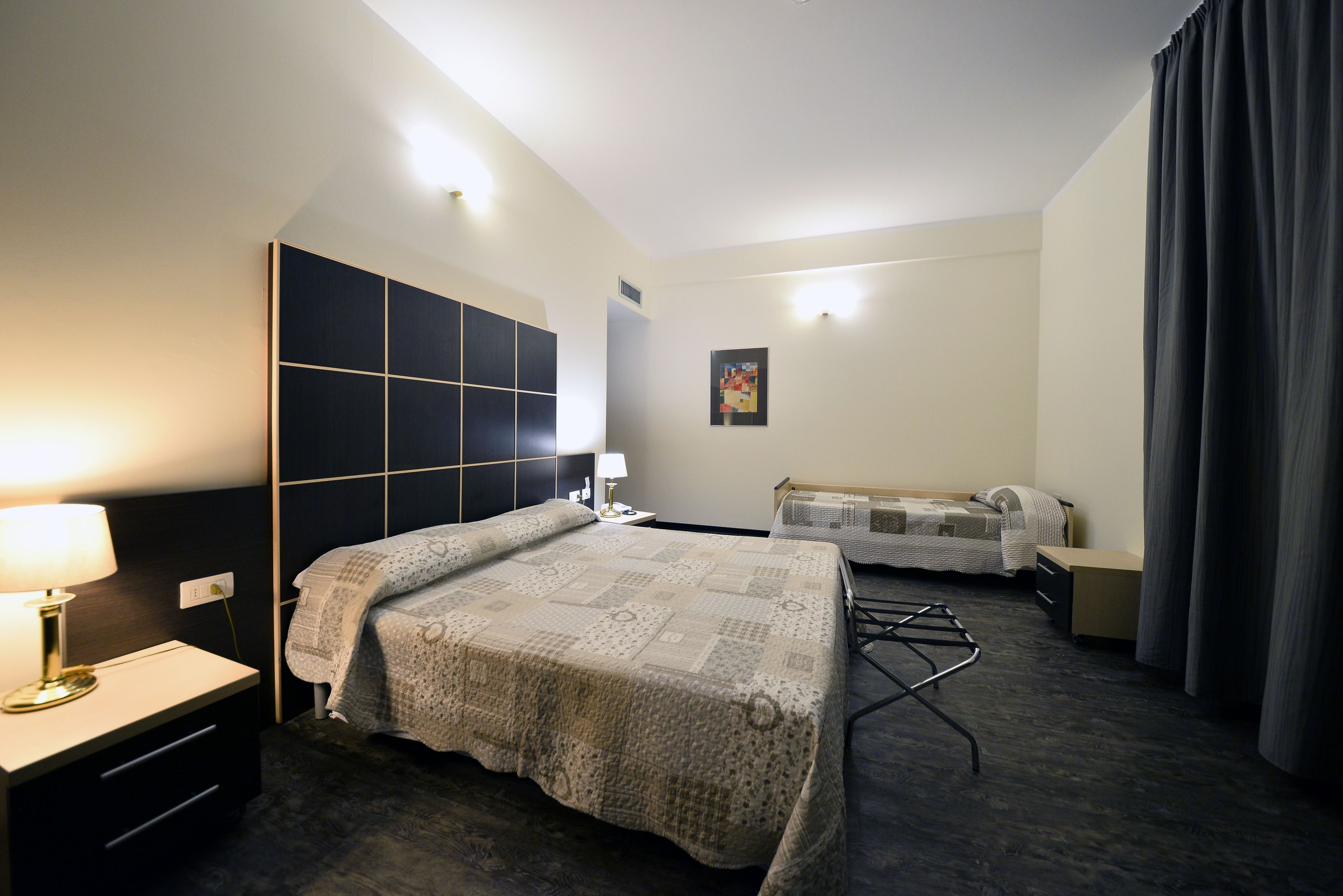 A hotel room with a large bed, a bedside table with a lamp, a small table with a lamp, a sofa, and a wall-mounted TV.