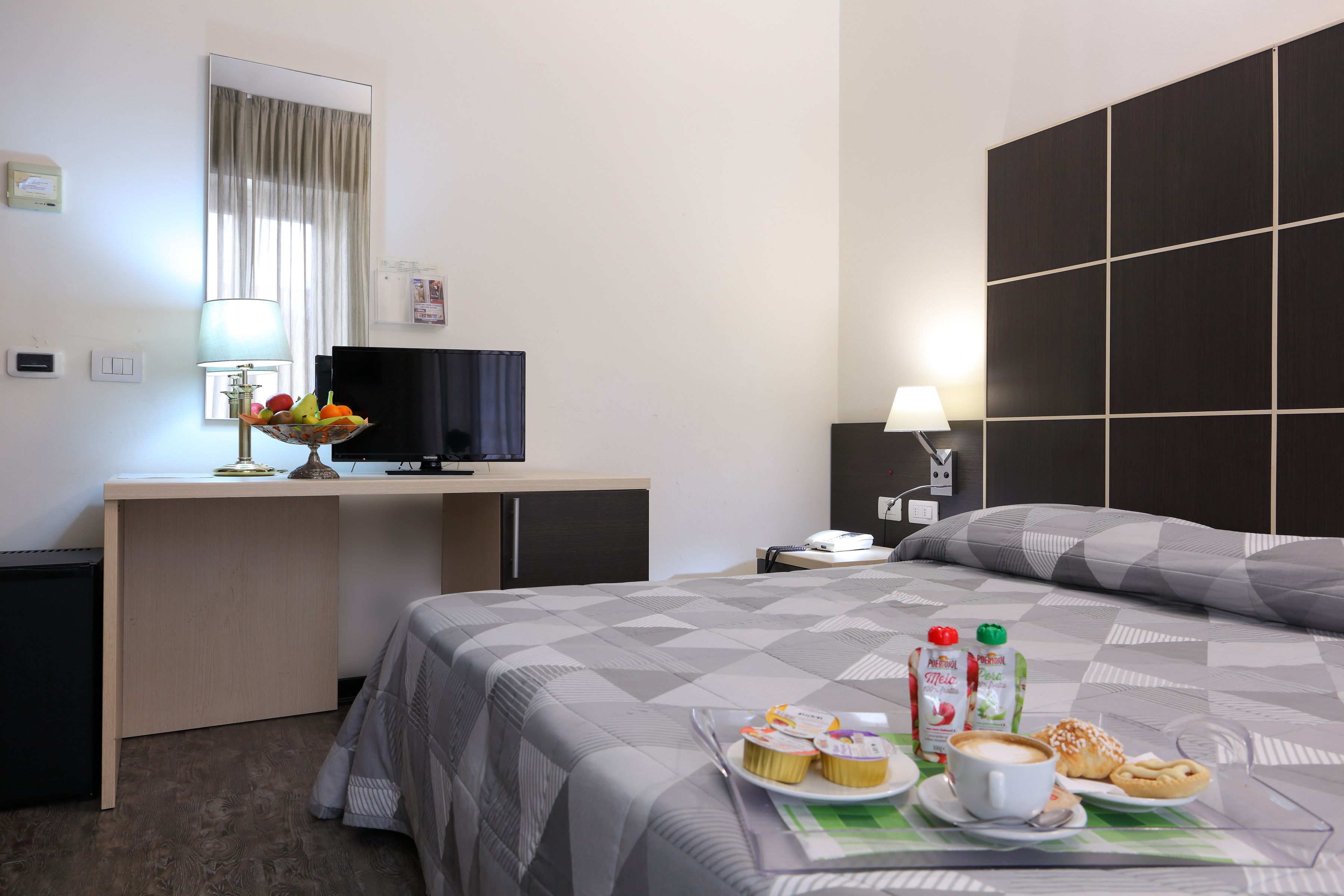 A hotel room with a bed, a desk with a television, a lamp, and a breakfast tray with pastries and beverages.