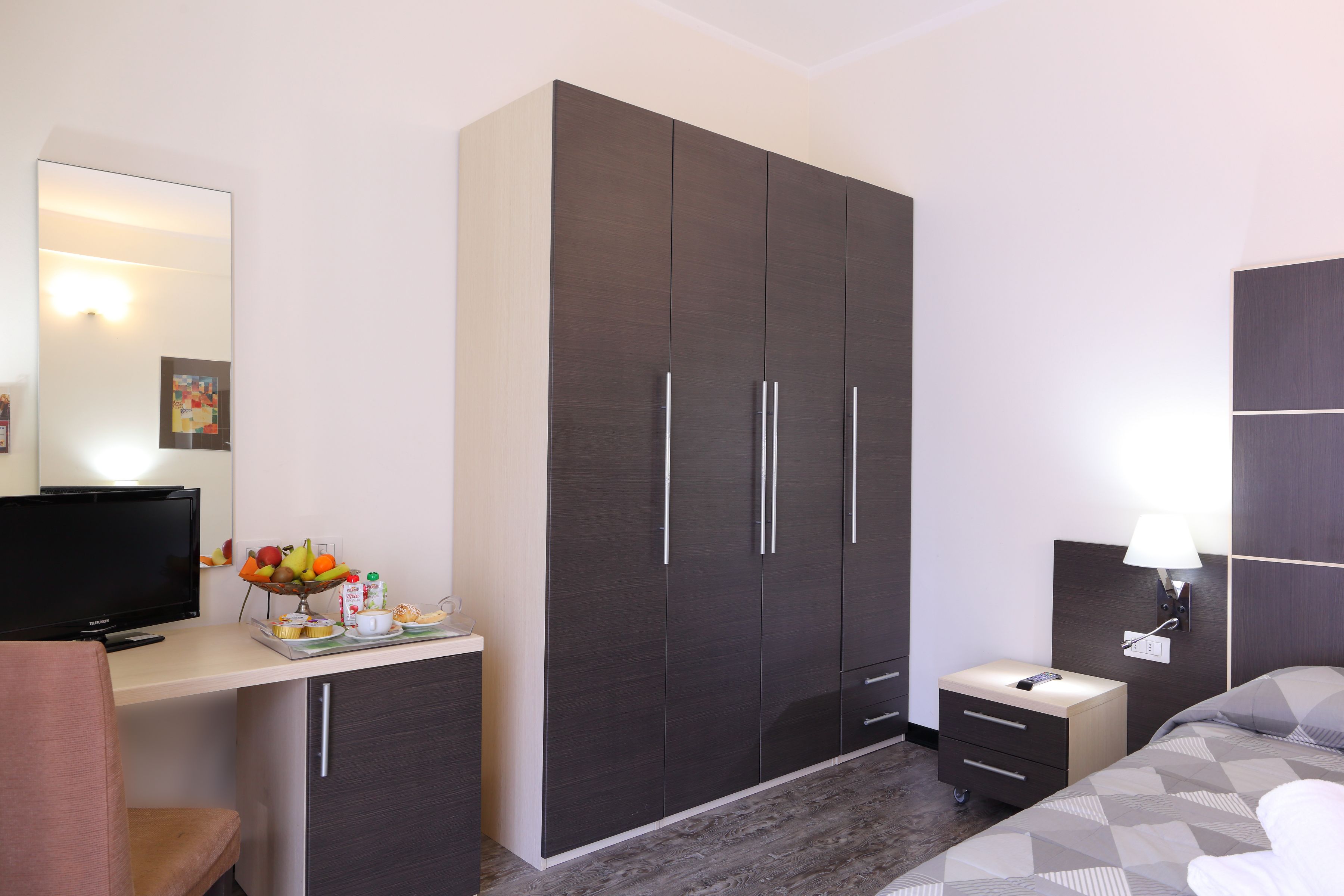 A modern hotel room with a large wardrobe, a desk with a flat-screen TV, a bedside table with a lamp, and a bed with a patterned bedspread.