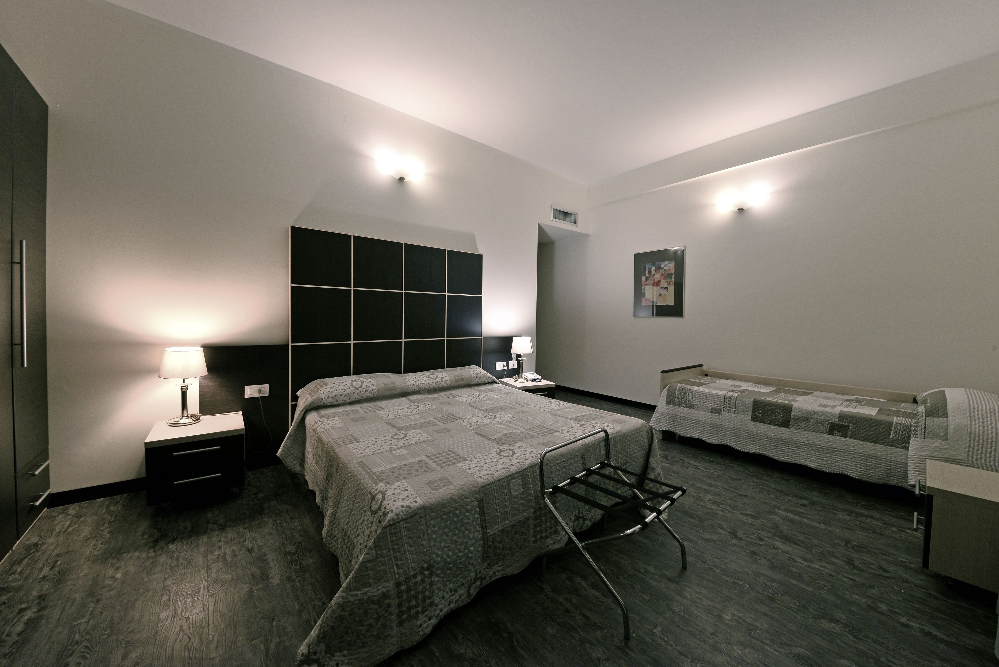 Una habitación de hotel moderna con una cama grande, una cama adicional, una mesita de noche con una lámpara, un televisor montado en la pared y un armario.