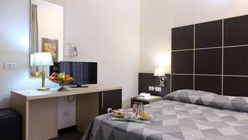 Camera d'albergo con un letto, una scrivania con lampada, una televisione e un vassoio colazione con frutta e bevanda.