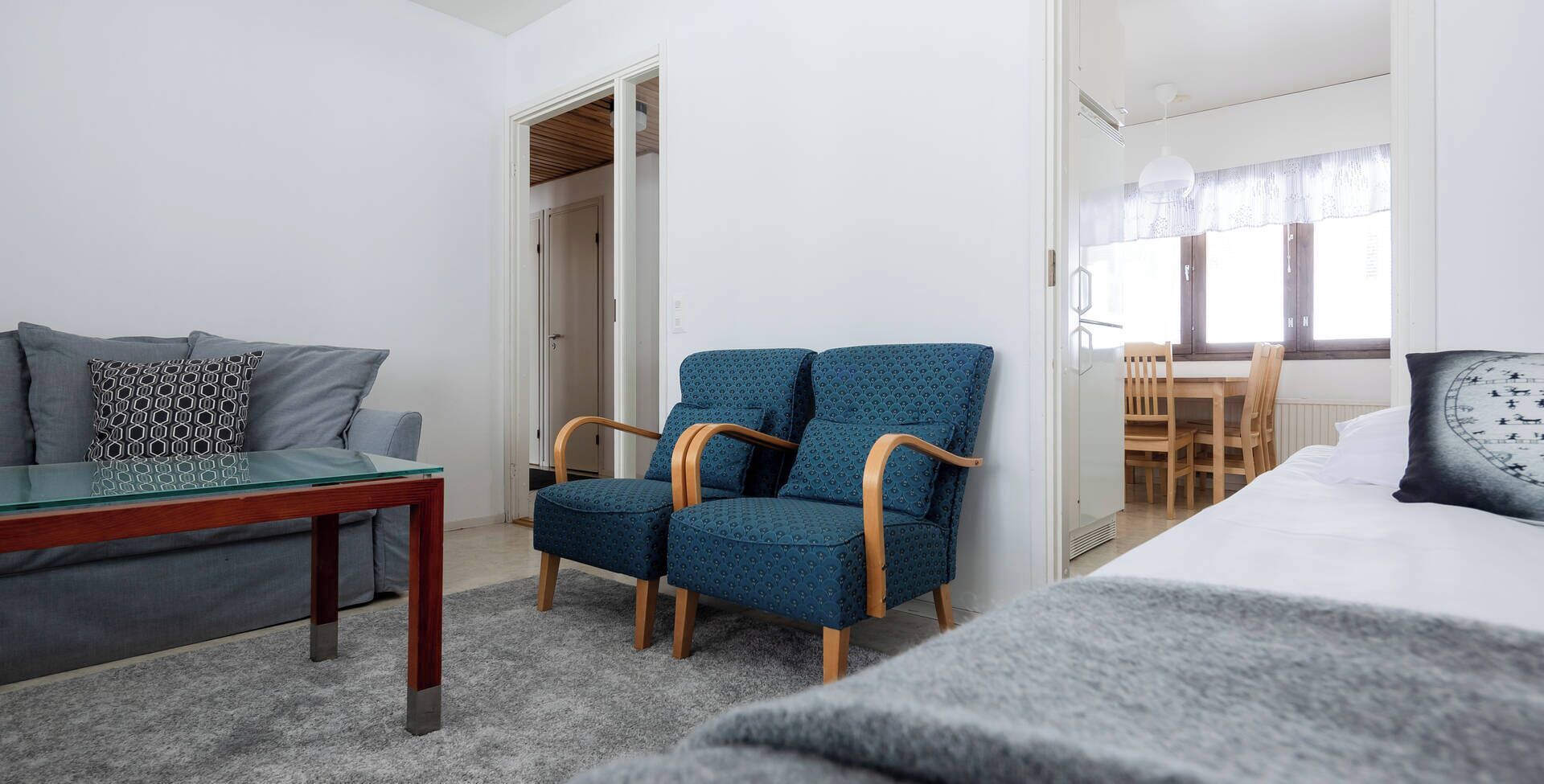 A modern hotel room with a grey sofa, a blue patterned armchair, a wooden side table, and a bed with a white headboard.