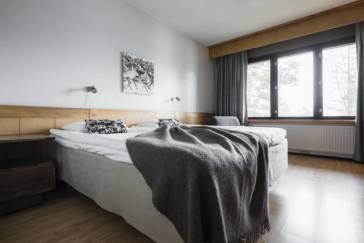A modern bedroom with a large bed, a nightstand, a window with curtains, and a wall-mounted light.