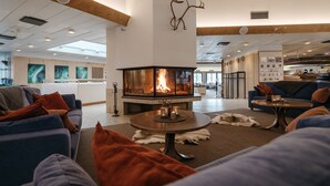 A cozy lounge area with a central fireplace, comfortable seating, and a view of the kitchen in the background. - Lapland Hotels Hetta (Enontekio)