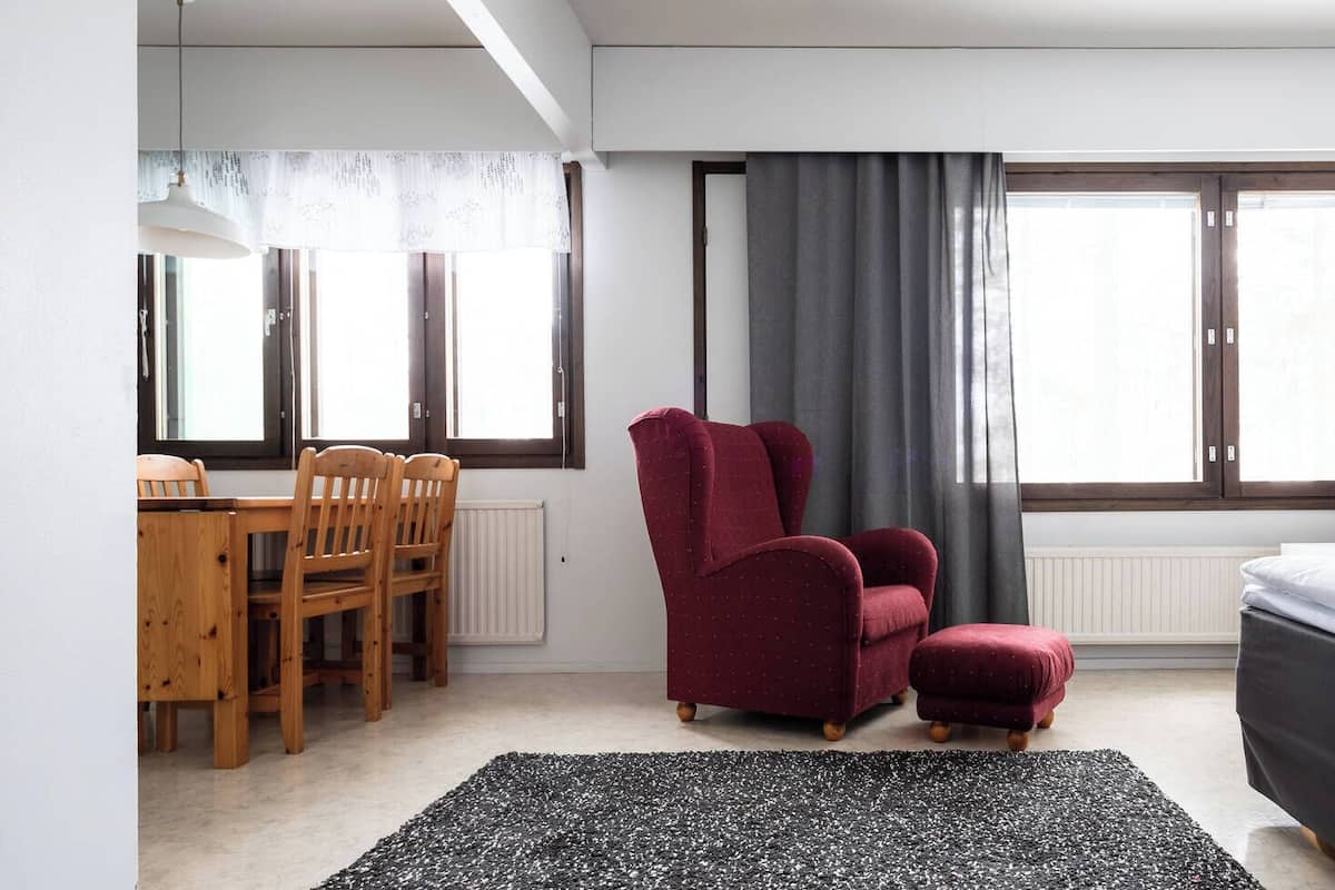 A modern hotel room with a wooden dining table, two red armchairs, a black and white rug, a bed, and a TV.