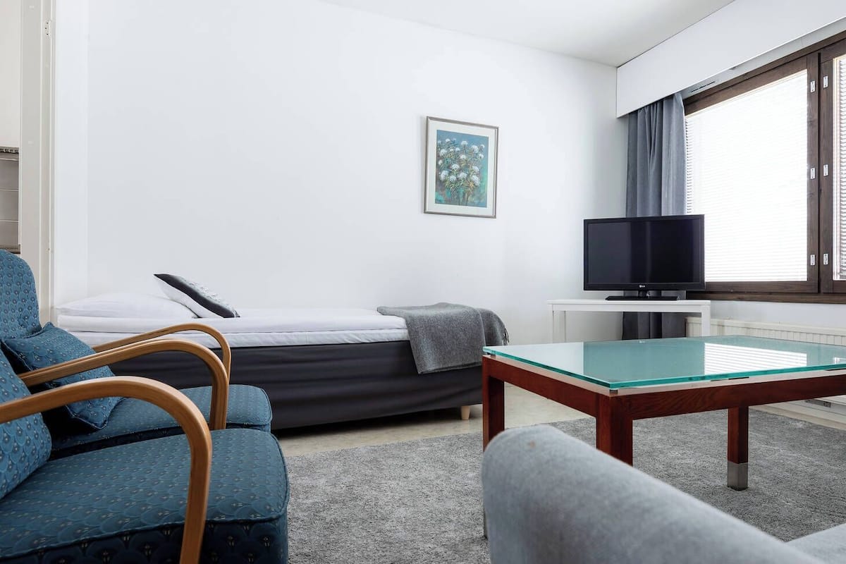 A modern, well-lit room with a bed, a blue armchair, a glass-top coffee table, a television, and a window with blinds.