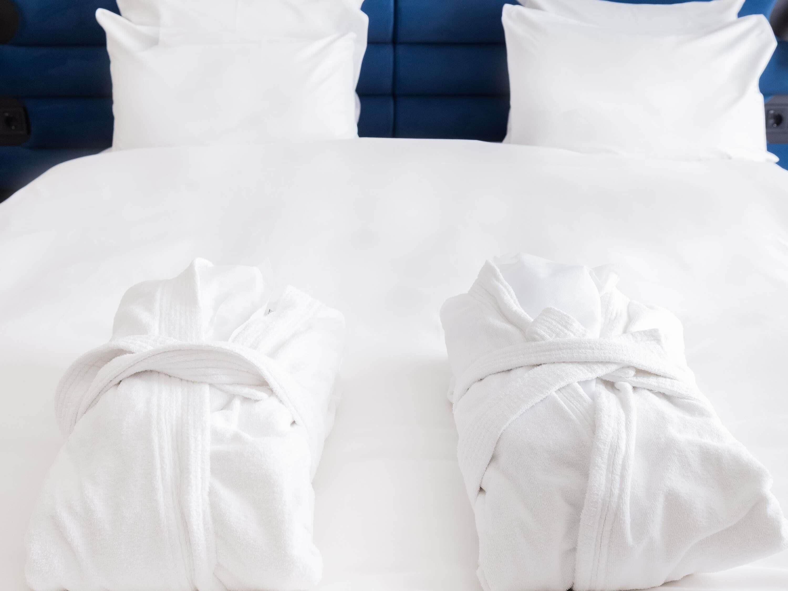 Una cama con sábanas blancas y dos batas blancas dobladas al pie.