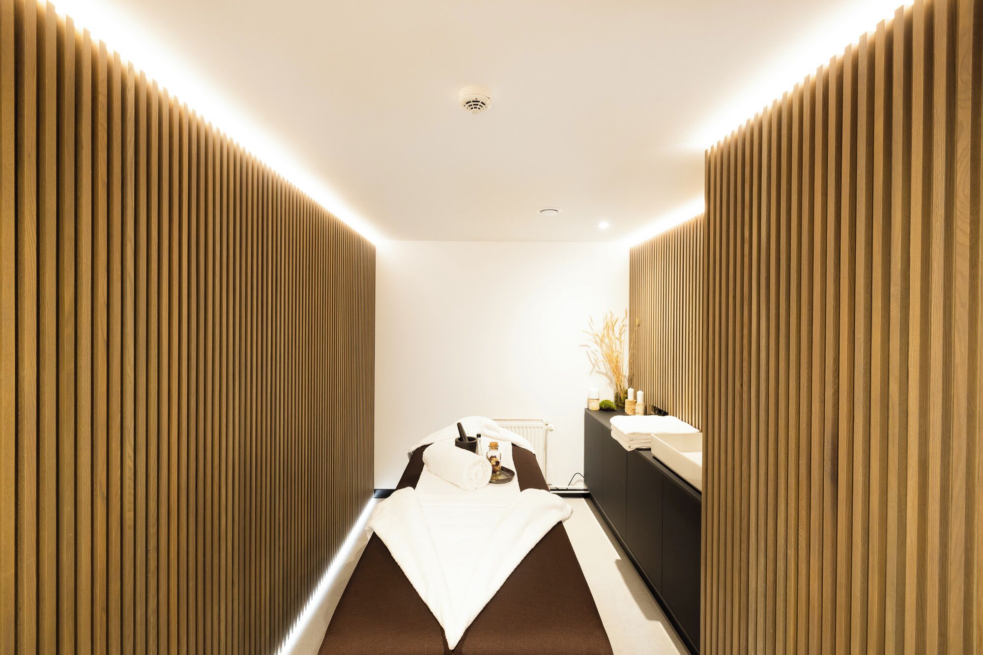 Moderni spa-huone, jossa on keskeisellä paikalla hierontapöytä, pieni meikkipöytä peilillä sekä seinälle asennettu lämmitin.