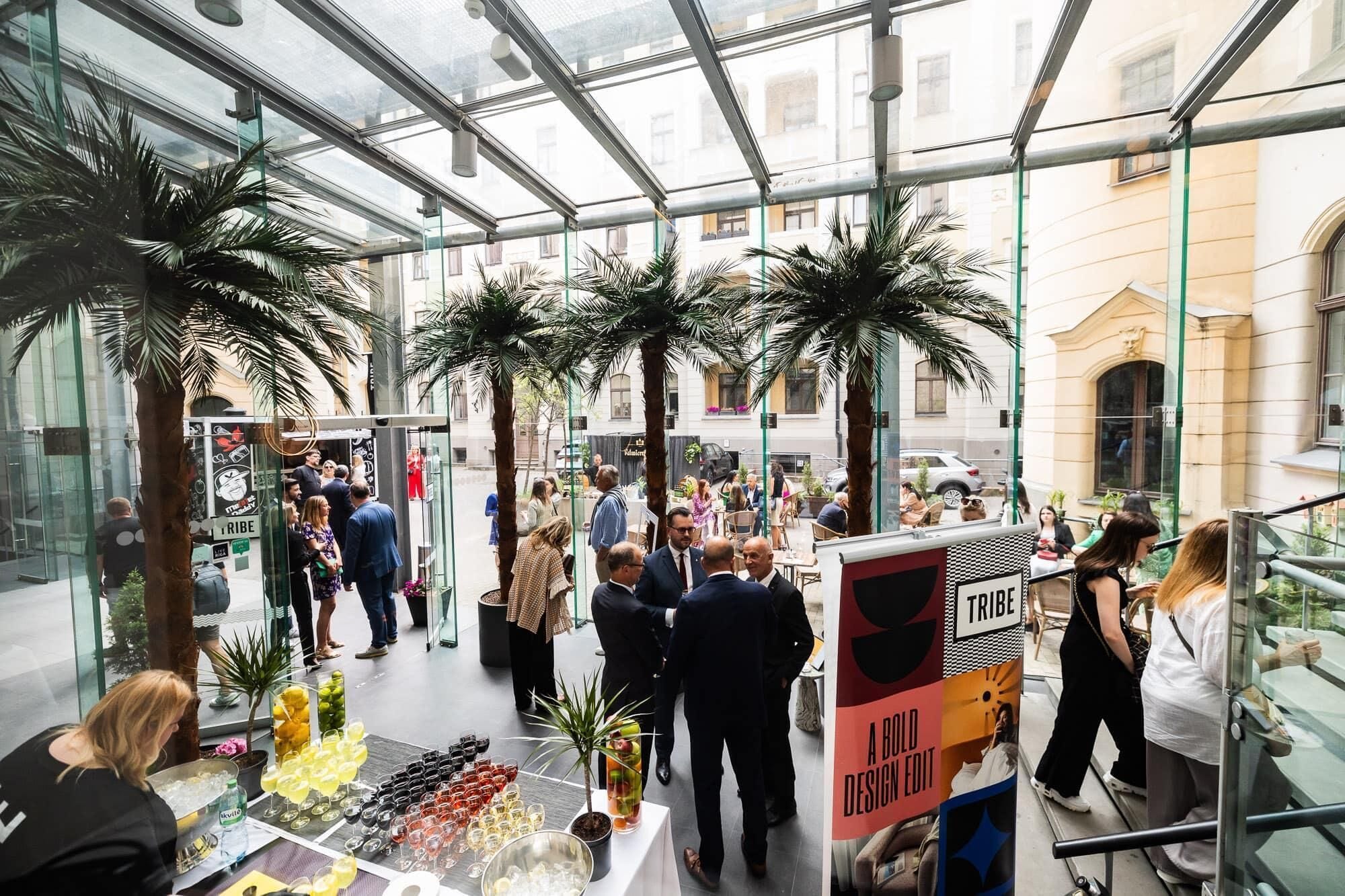 A glass-enclosed atrium with palm trees, a crowd of people, and a sign reading 'A BOLD DESIGN EDIT'.
