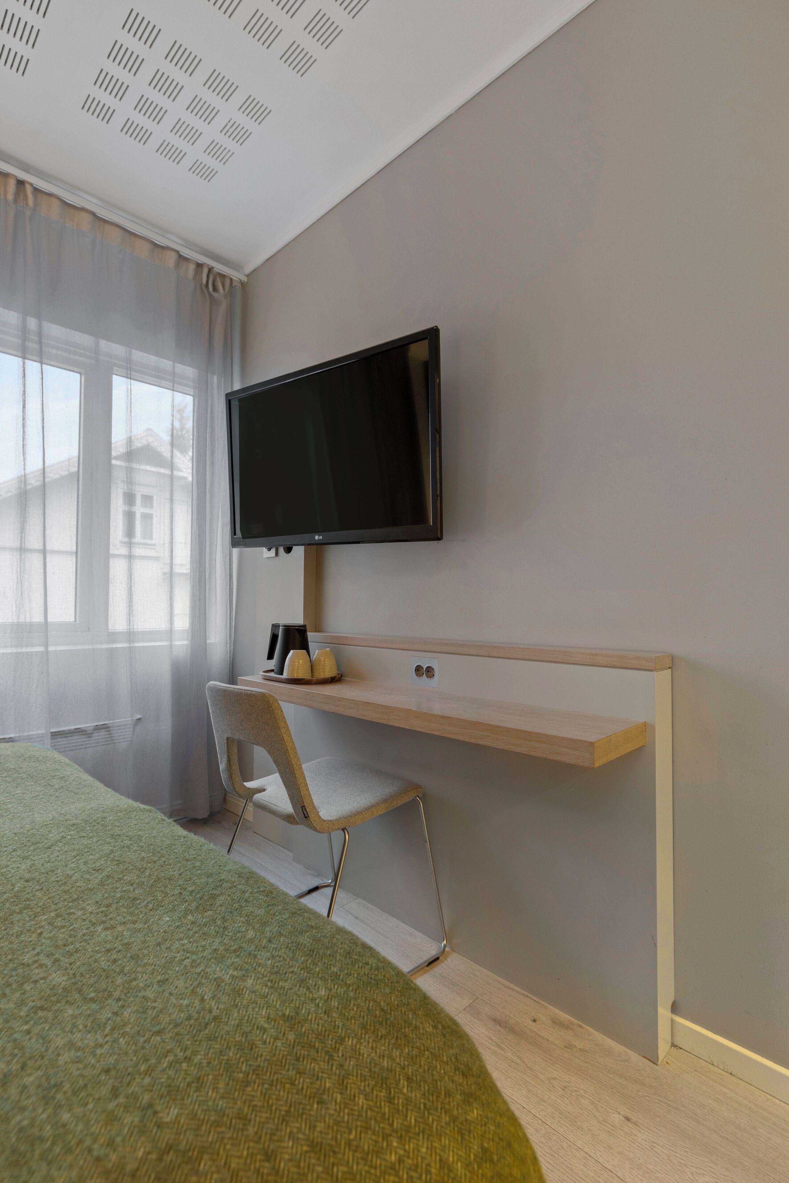 A hotel room with a bed, a desk with a computer, a chair, a TV mounted on the wall, and a window with sheer curtains.