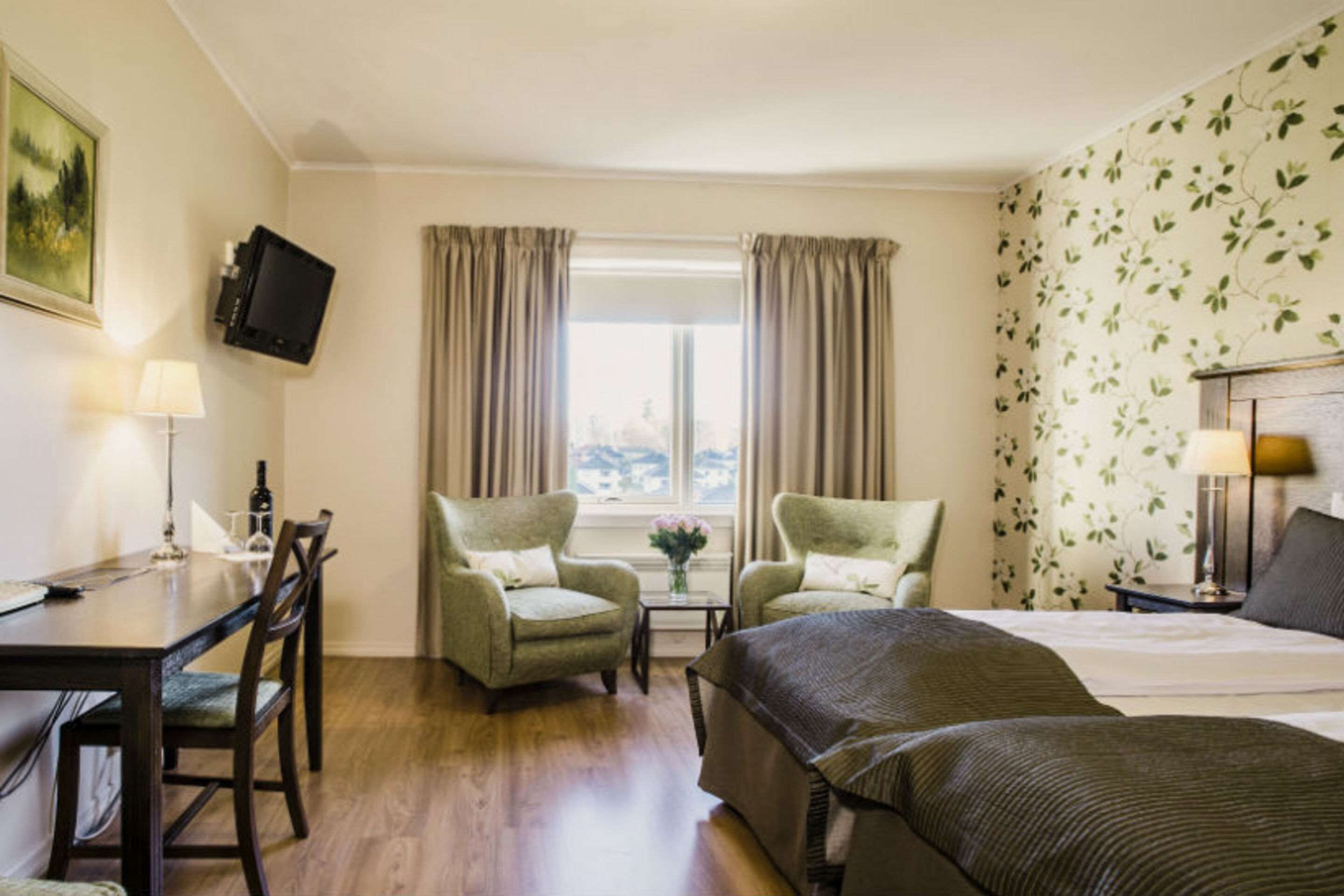 A hotel room with a bed, two armchairs, a small table with a vase, a television mounted on the wall, and a window with curtains.