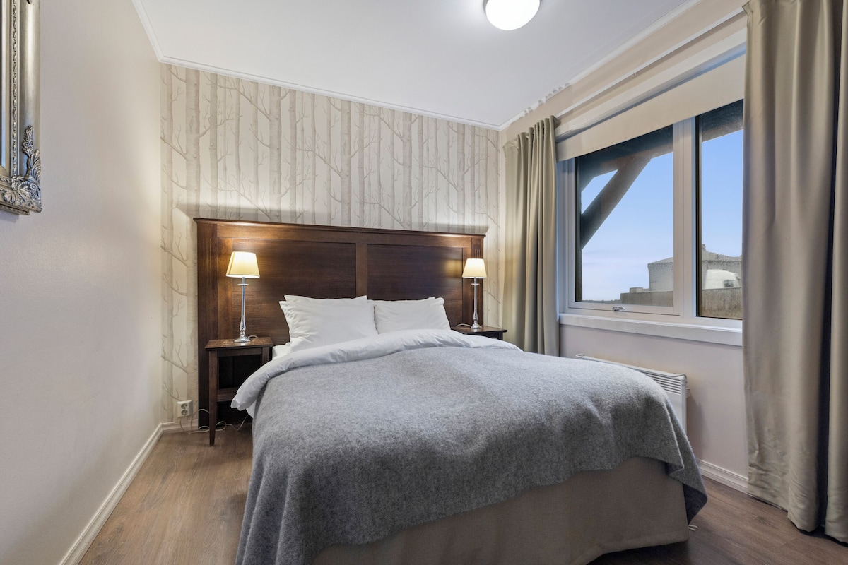 A hotel room with a large bed, two bedside tables with lamps, a window with curtains, and a view of buildings outside.