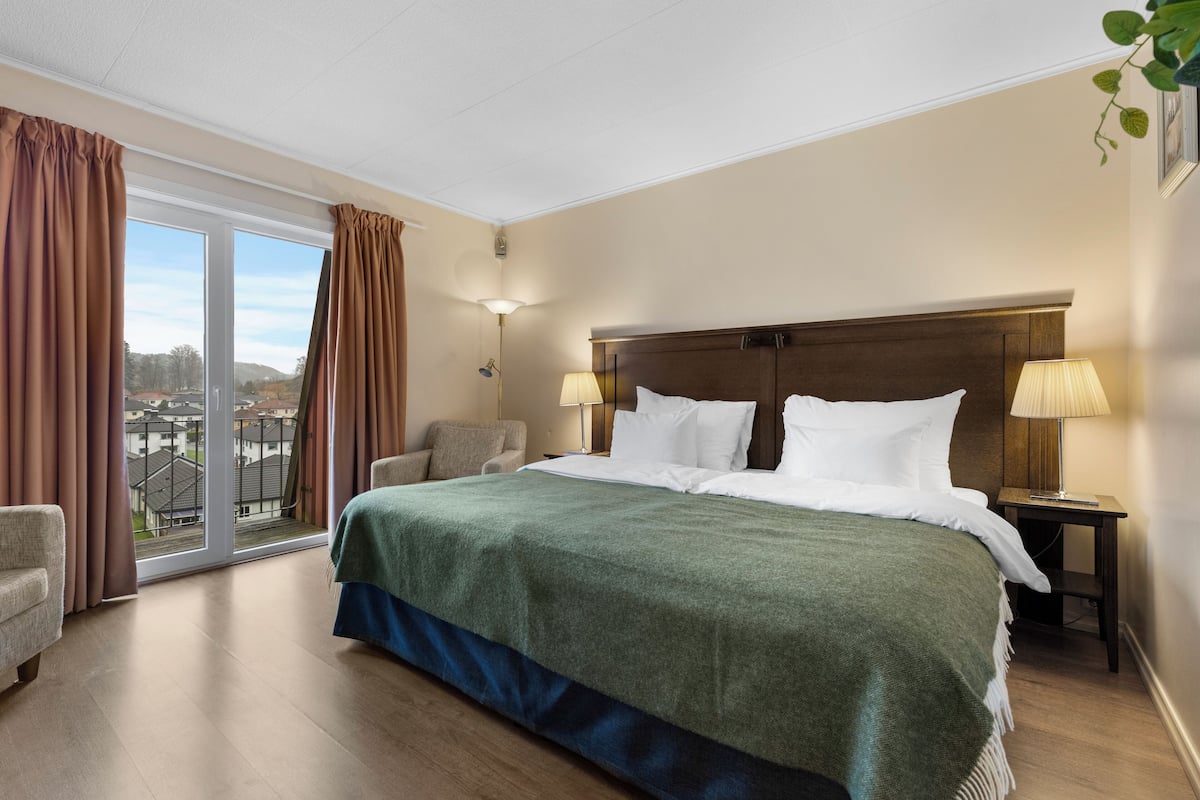 A hotel room with a large bed, two bedside tables with lamps, a chair, and a view of the outside.