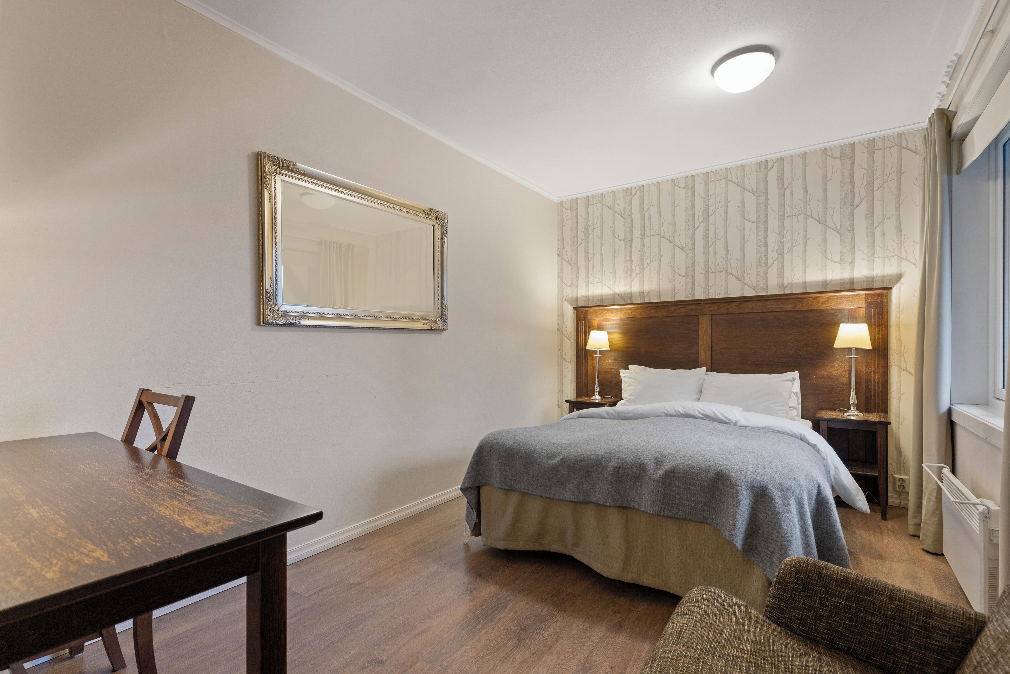 Quarto de hotel com uma cama de madeira, uma mesa de cabeceira com luminárias, uma cadeira e um espelho grande na parede.