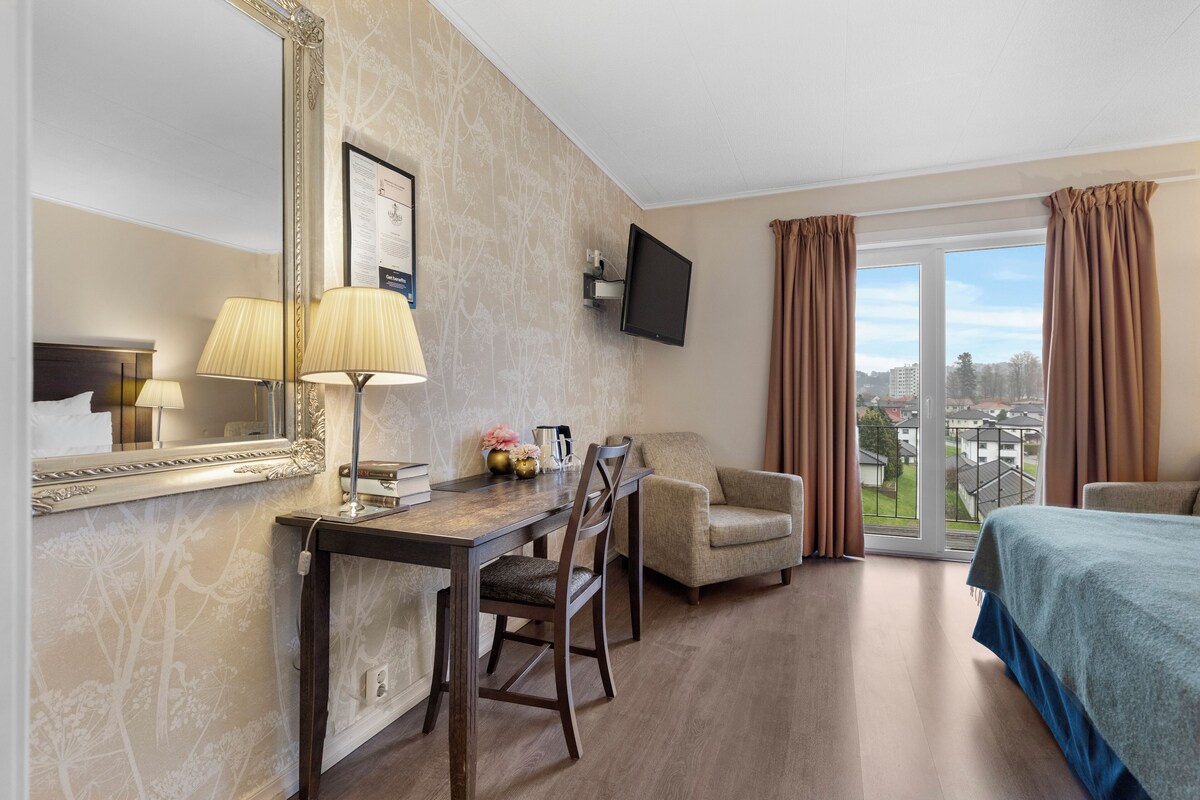 A hotel room with a bed, a desk with a chair, a television, and a view of the city through the window.